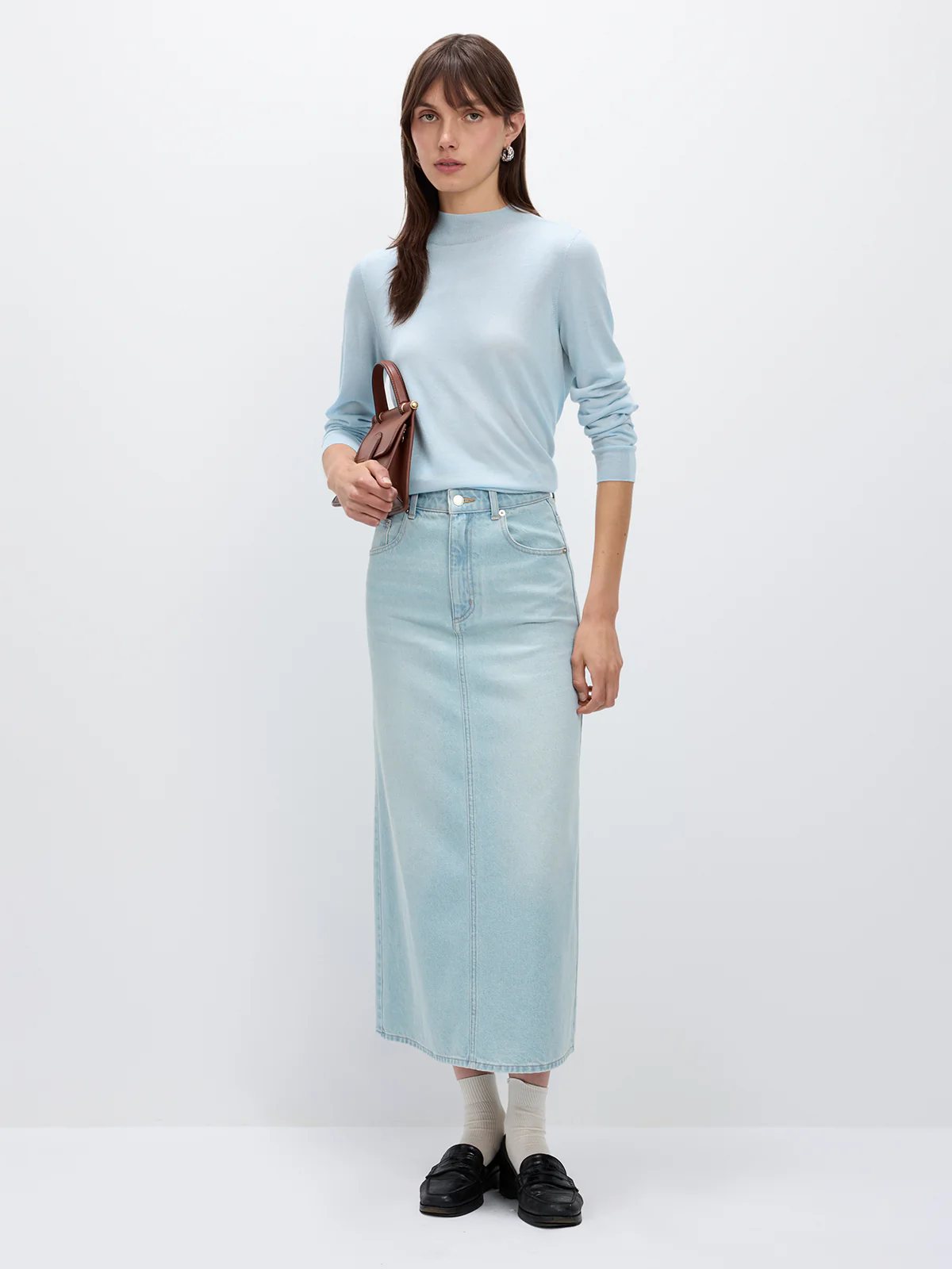 Model wearing a sage green silk lyocell mock-neck top with long sleeves, holding a woven clutch, paired with a skirt and sandals.