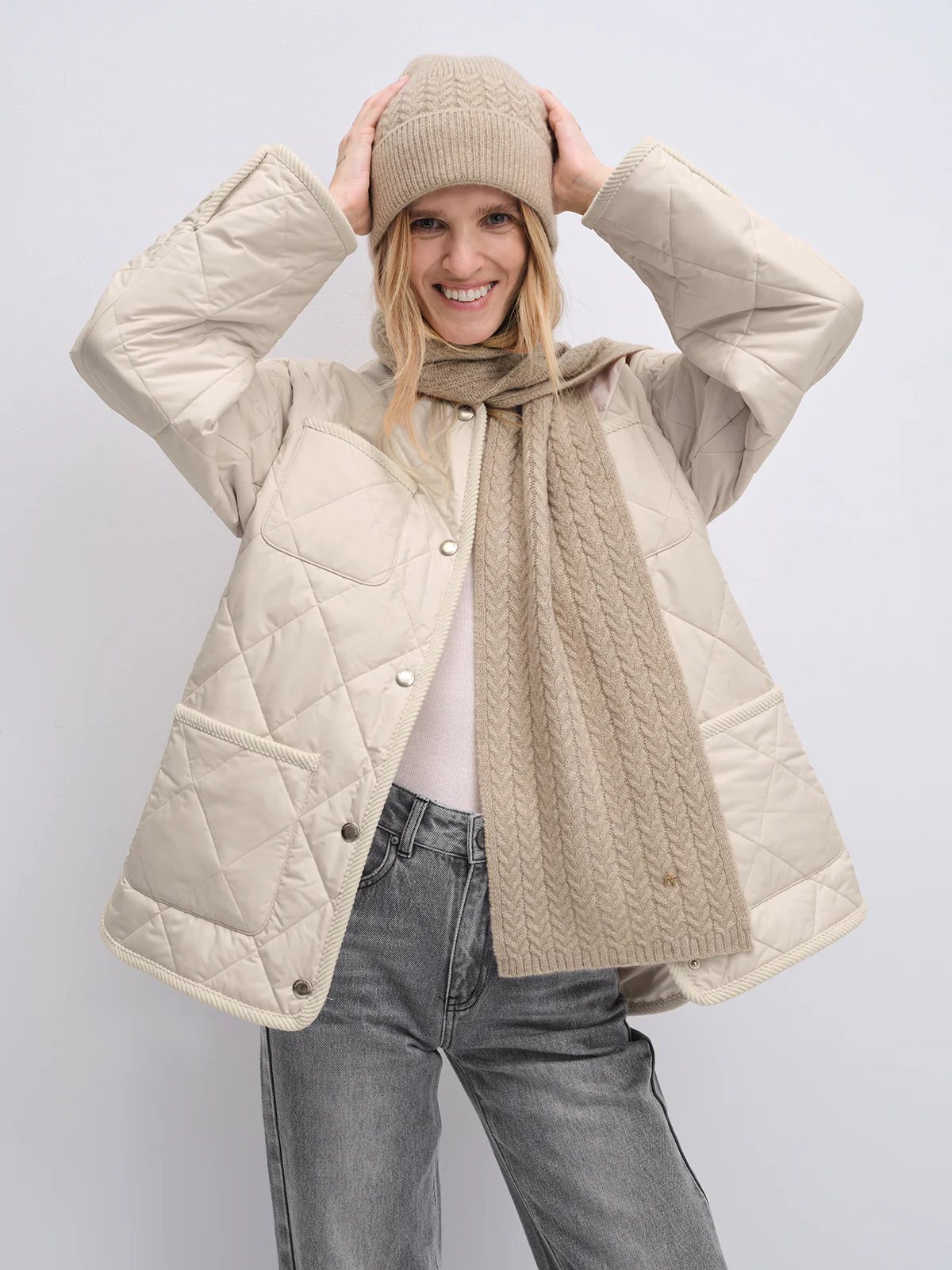 Model in off-white quilted textured jacket with rope-trimmed collar and cuffs, wearing matching beanie and scarf, hands adjusting hat.