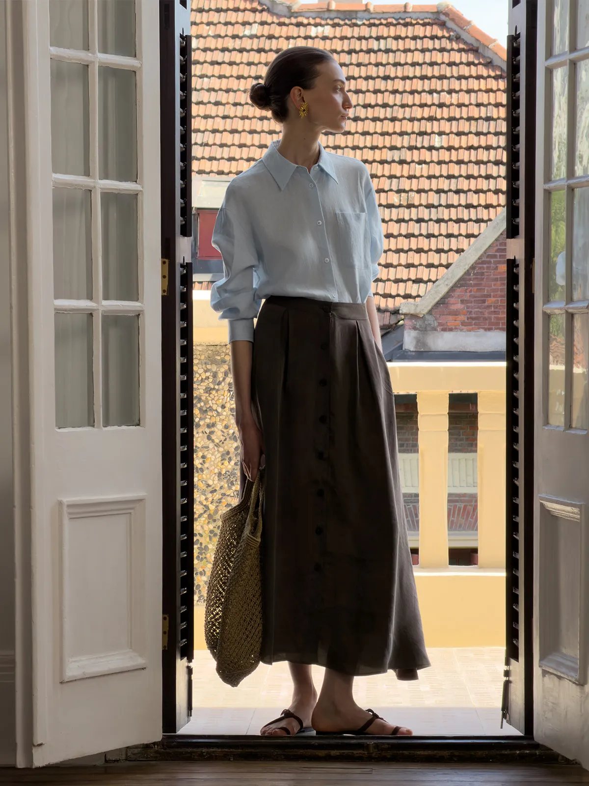 Model standing in doorway wearing light blue linen shirt and brown midi skirt, overlooking rooftop tiles