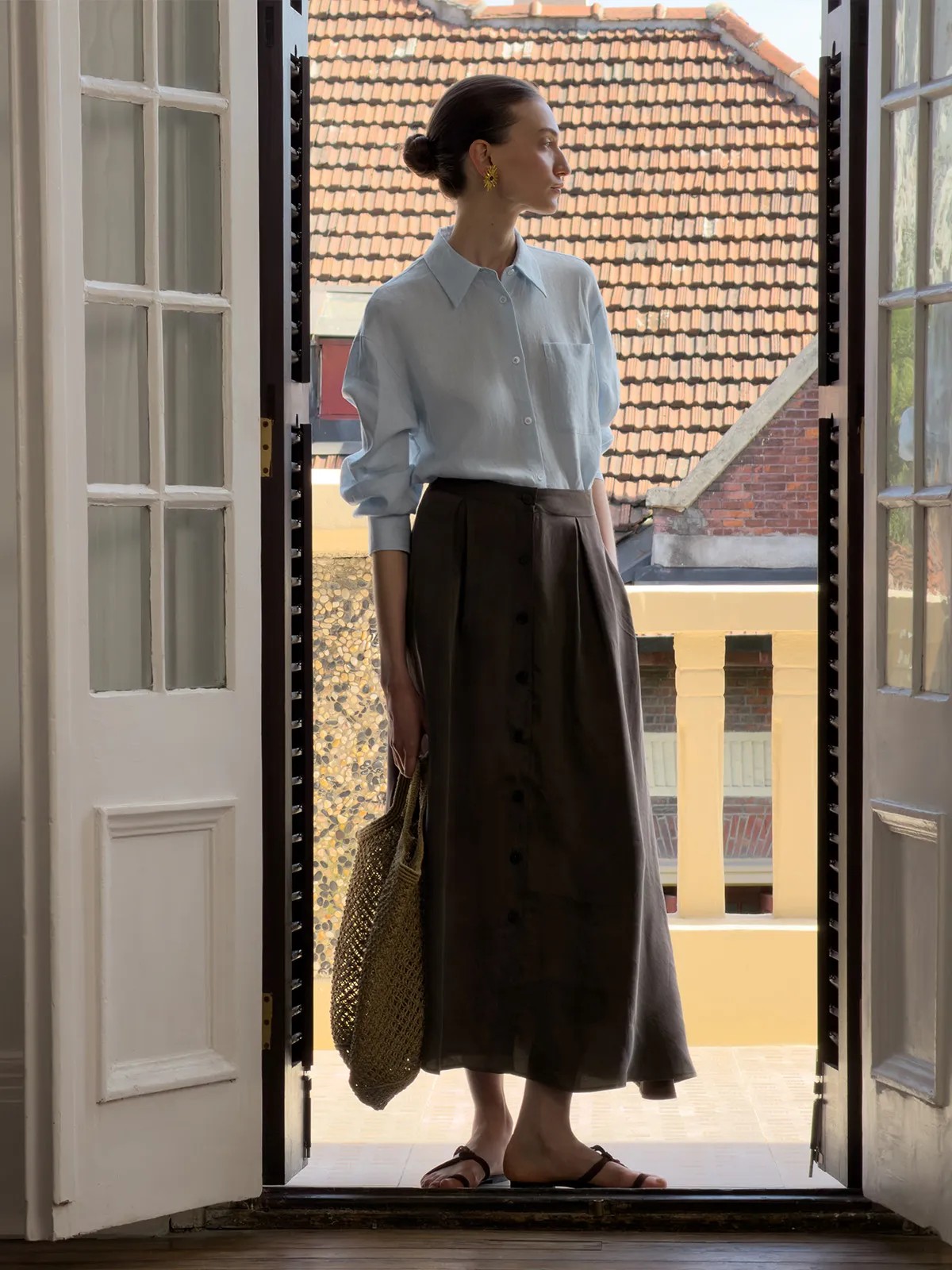 Model standing in doorway wearing light blue linen shirt and brown midi skirt, overlooking rooftop tiles