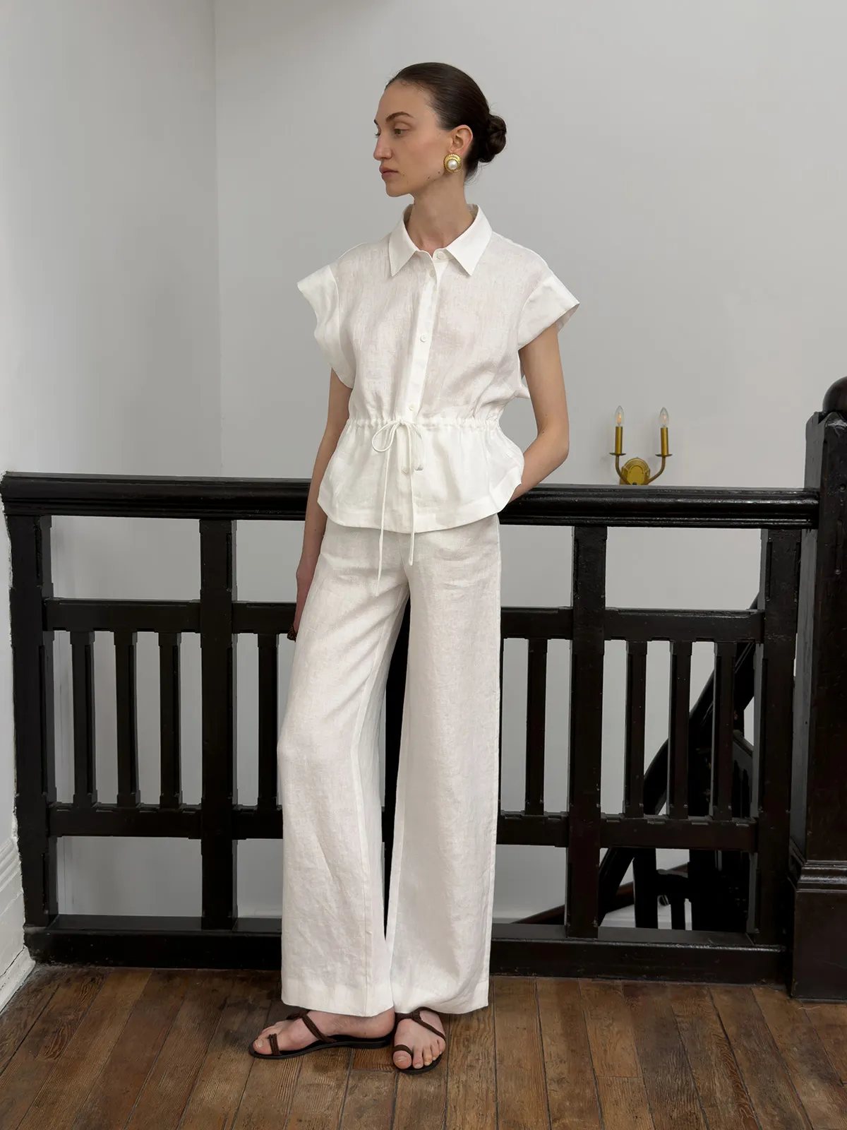 Side profile of model in ivory CLORIS linen trousers and white peplum shirt, leaning against black railing — minimalist elegance