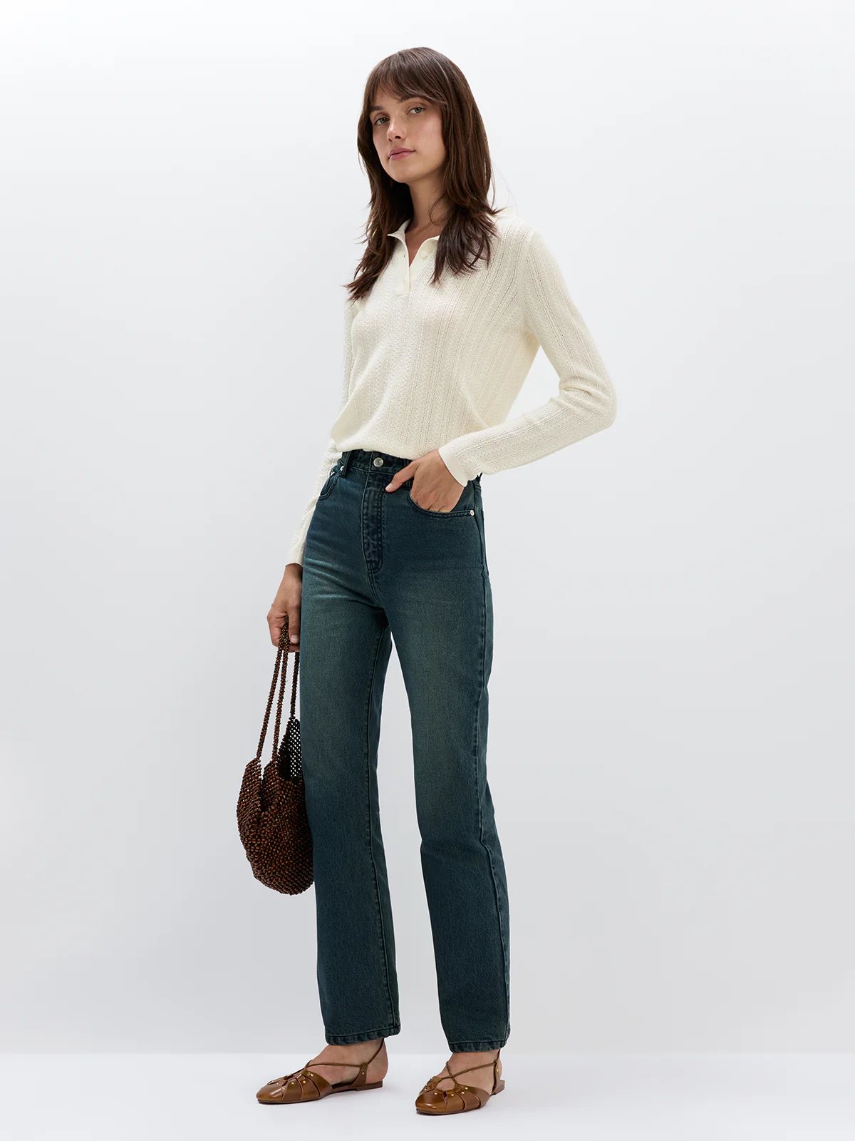 Side view of model wearing Blue mid-rise slim straight jeans with a sweater and holding a woven bag.
