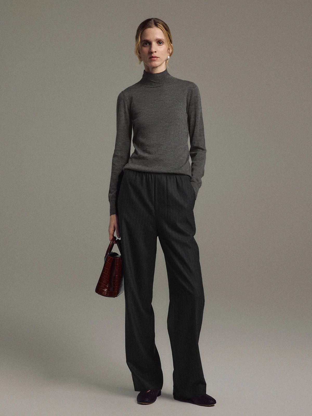 Model wearing Gray/White pinstripe wool-blend straight trousers with elastic waist, paired with a gray turtleneck and holding a bag.