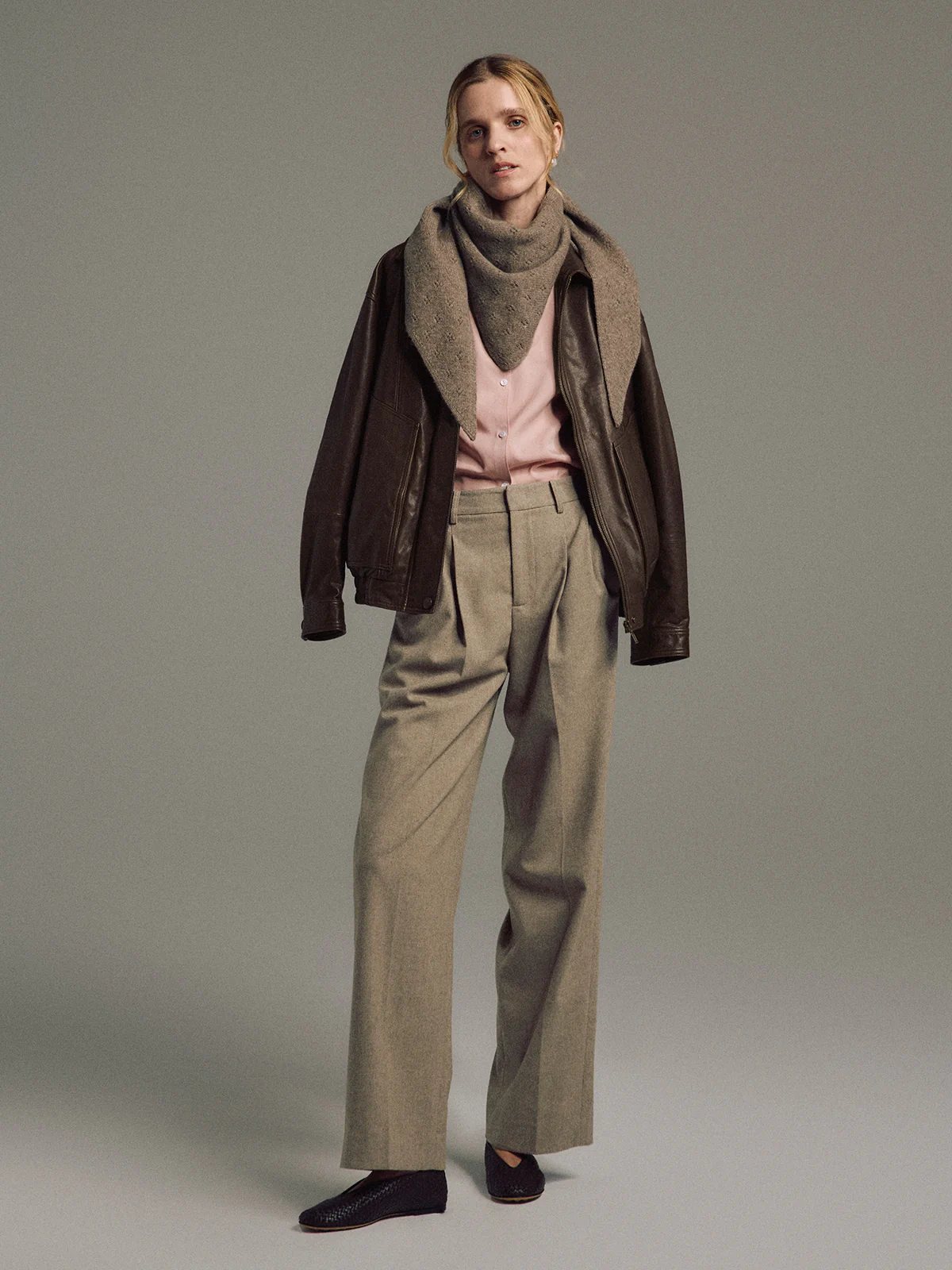 Model wearing Tan wool blend double-pleat straight-leg trousers layered under shirt and jacket, holding bag.