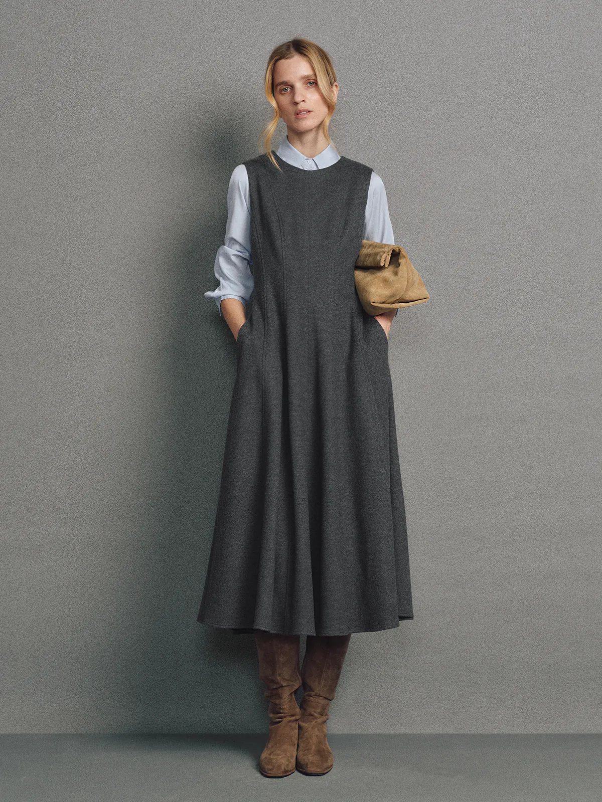 Side view of model in a sleeveless dark gray wool knit A-line dress layered over a light shirt, holding a clutch.