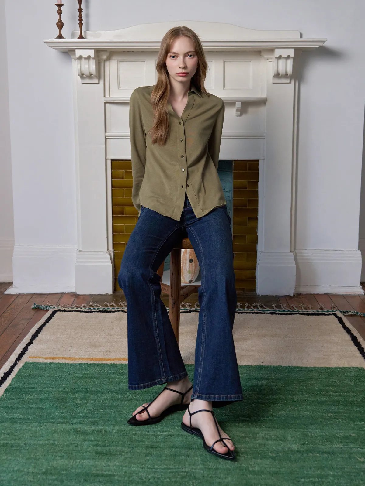 Model seated on wooden stool wearing olive COLETTE pique shirt with flared dark denim jeans, fireplace background