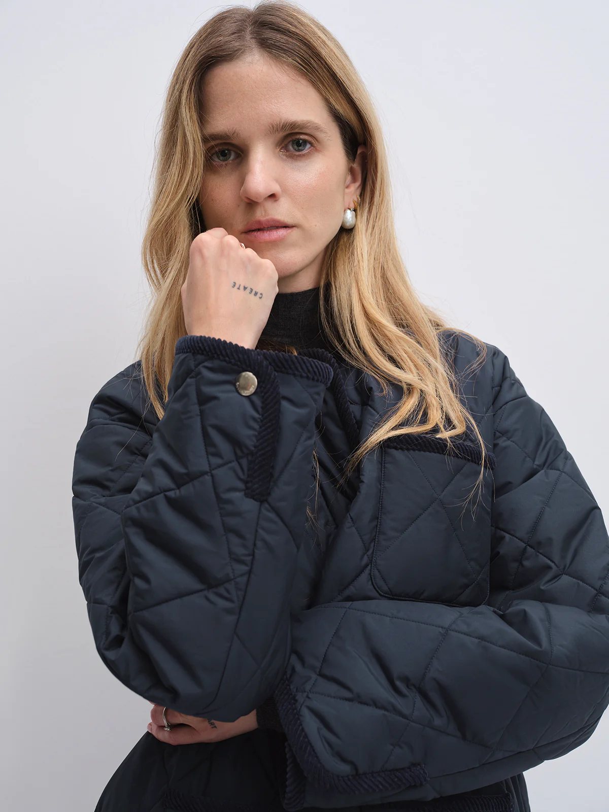 Close-up of model wearing navy quilted textured jacket with rope trim at collar and cuffs, hand near face showing wrist tattoo.