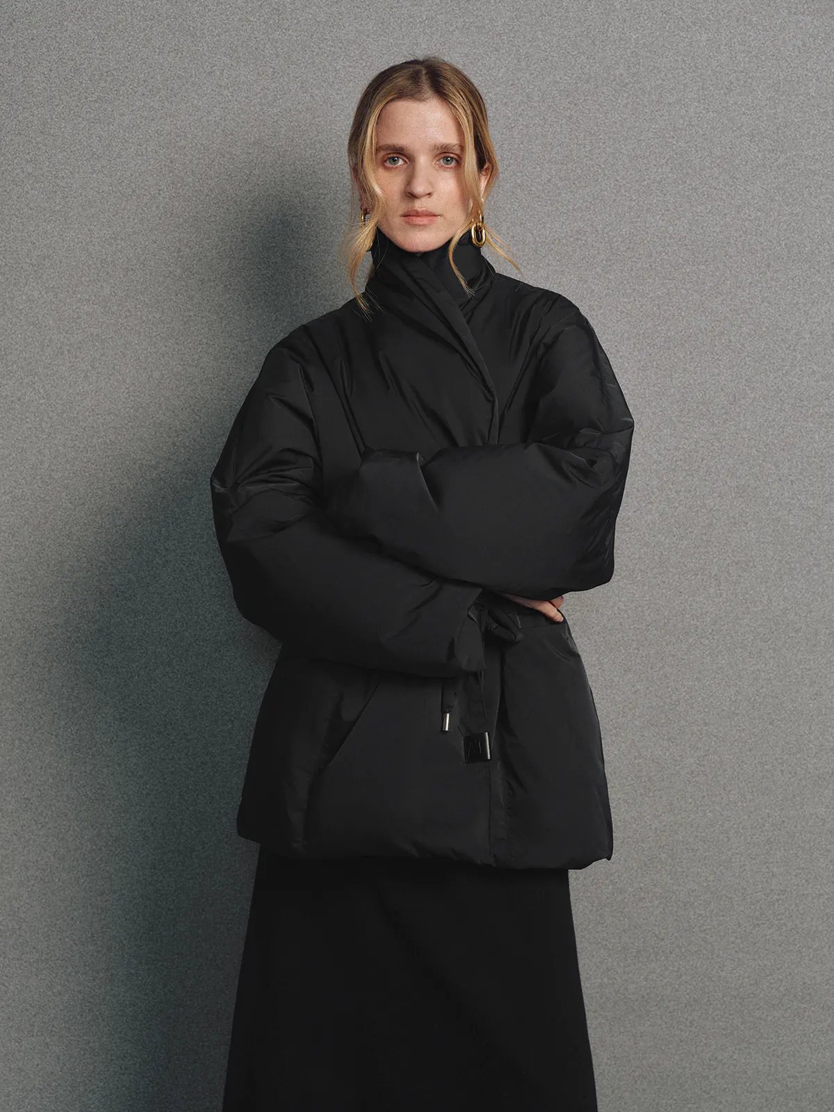 Front view of the Black OPHELIA relaxed wrap-style down coat with high collar and tie waist, arms crossed.