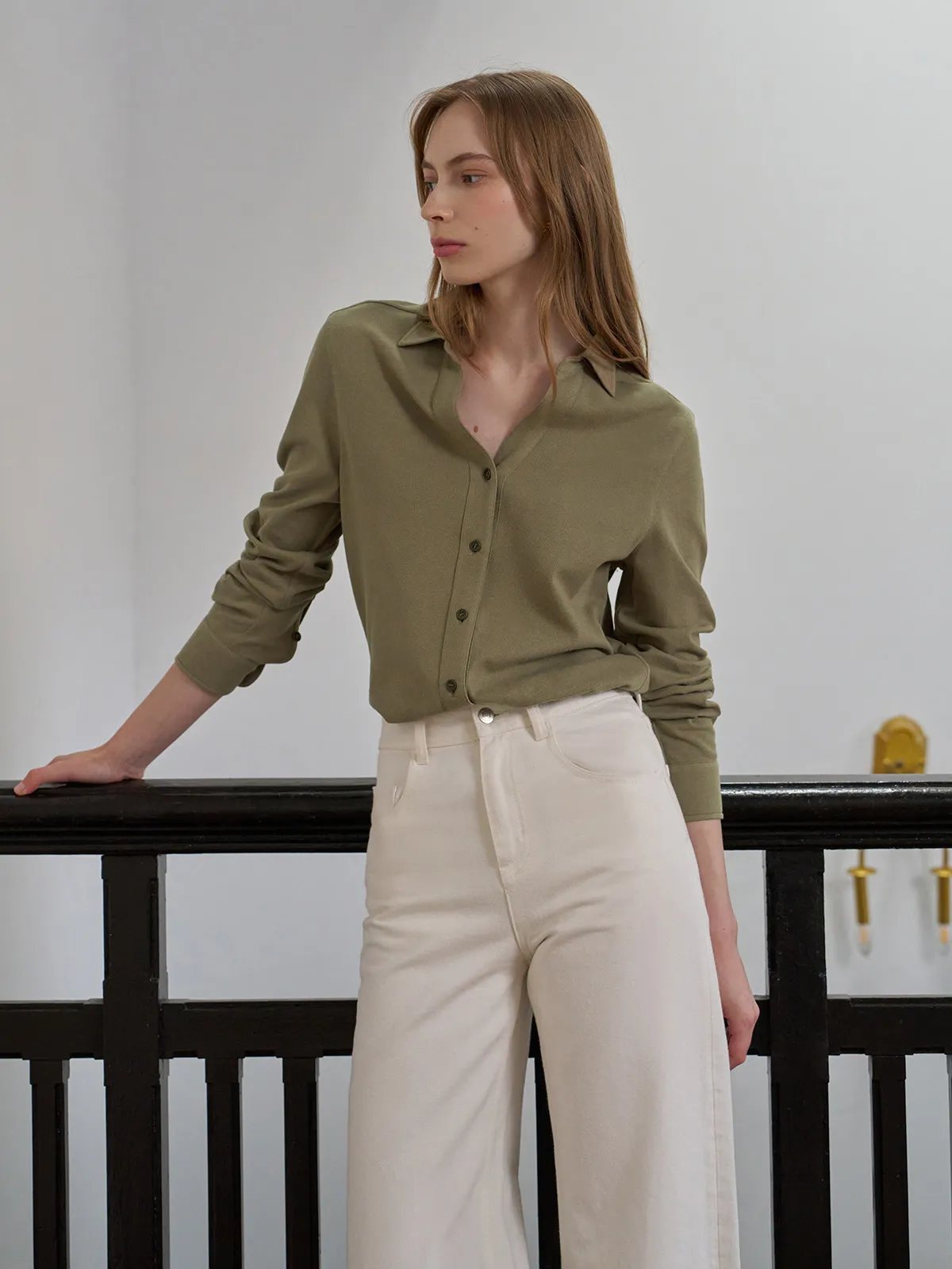 Side profile of model in olive COLETTE shirt and cream wide-leg pants, leaning on black railing, soft lighting