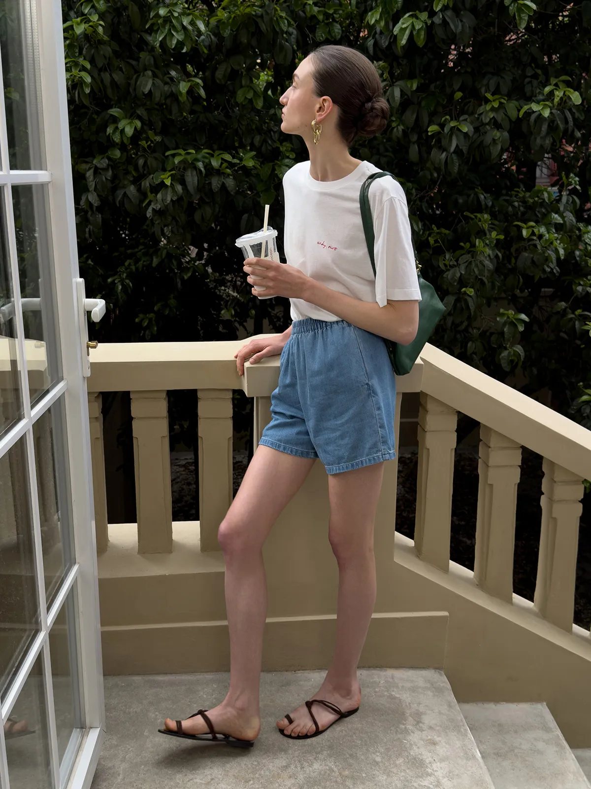 Model on balcony wearing WAYLON denim shorts, white tee, green bag, holding iced drink, lush greenery background