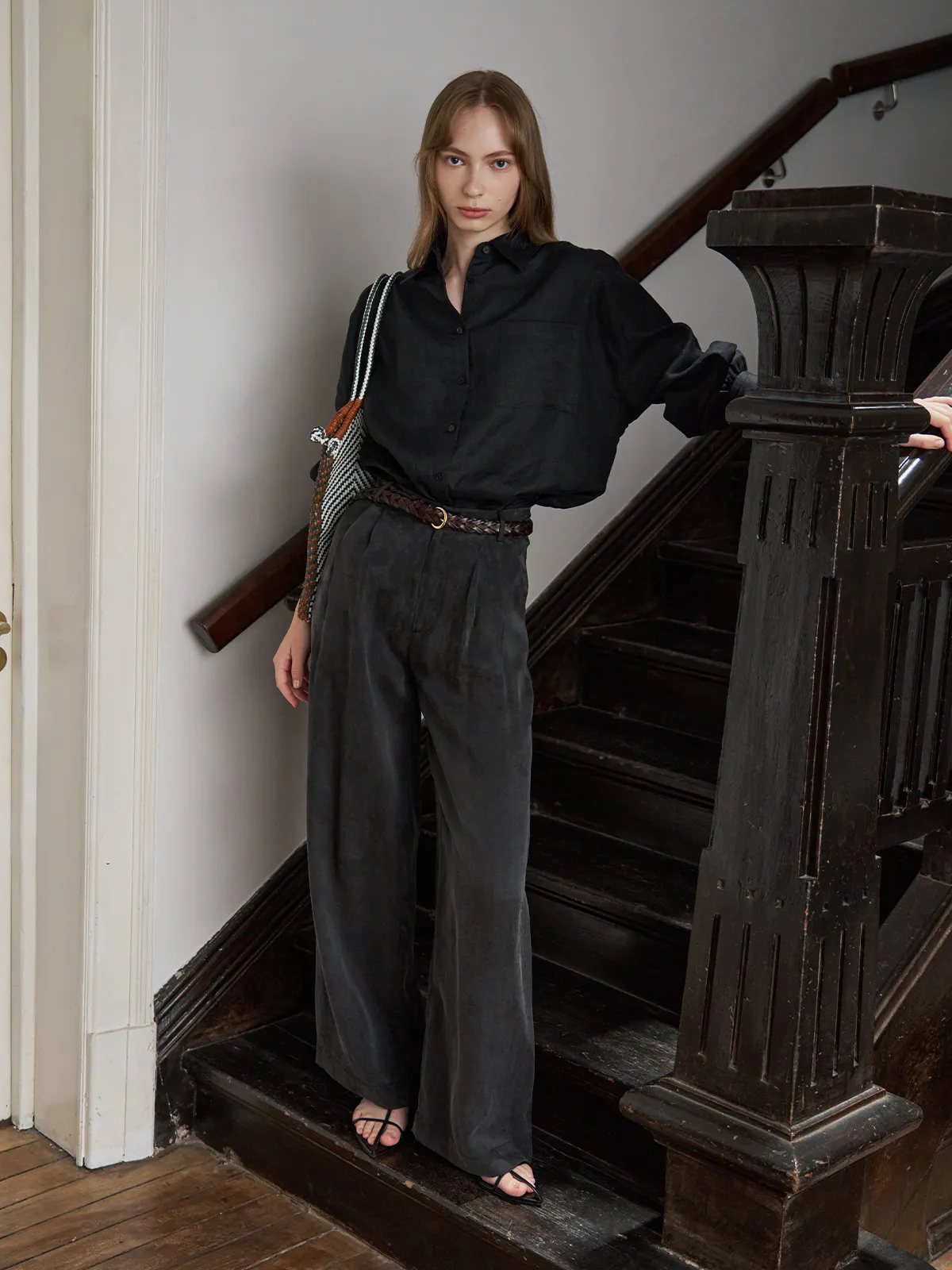 Model wearing Andreatang PLAIT woven leather belt with black shirt and wide-leg trousers on vintage staircase
