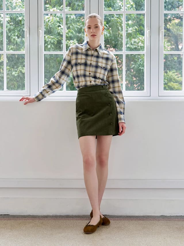 Model wearing Andreatang YUKI olive corduroy A-line skirt with plaid shirt, minimalist styling against white window backdrop