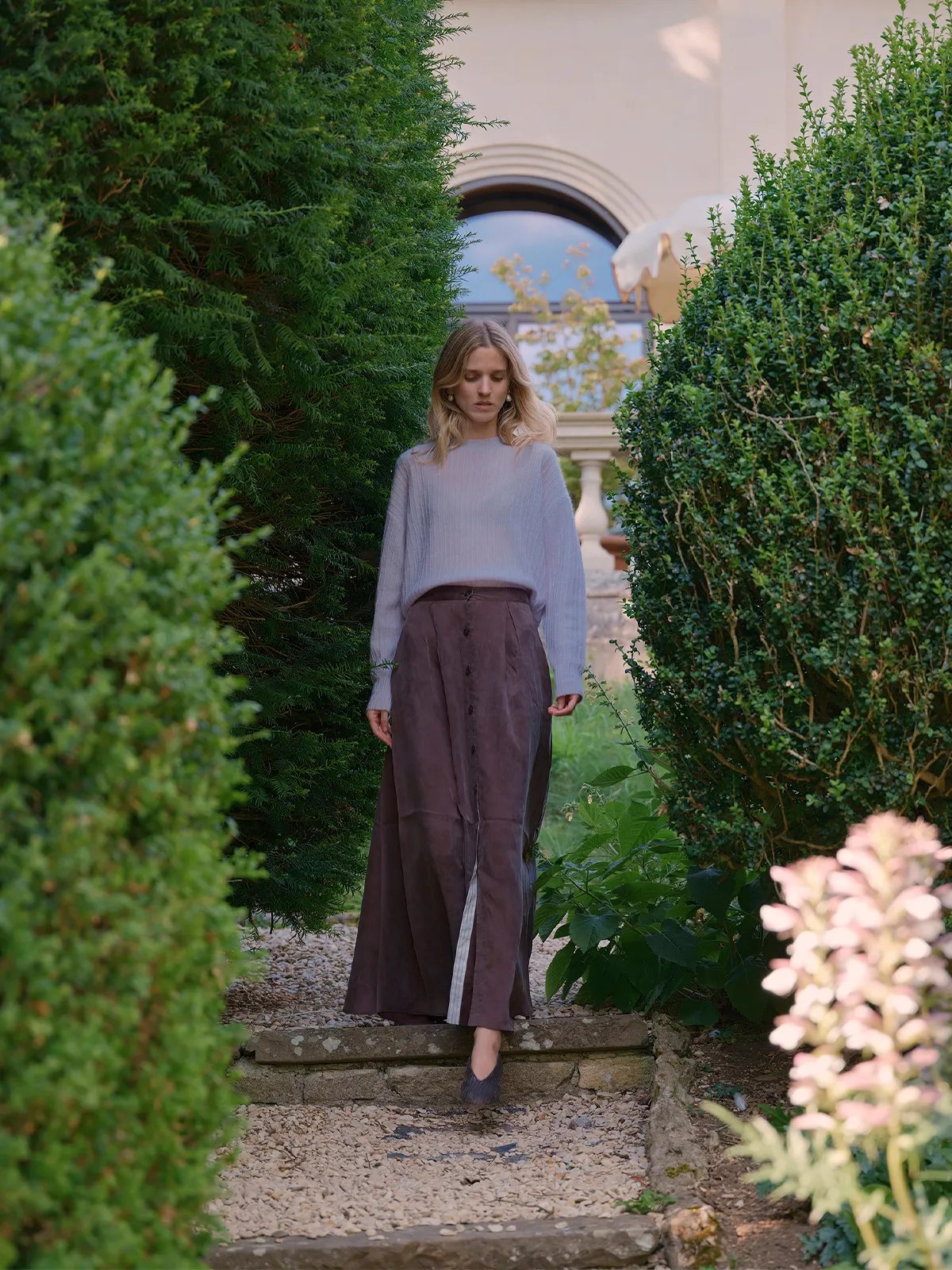 Model wearing lavender ELFIE jumper with brown button-front skirt in serene garden pathway
