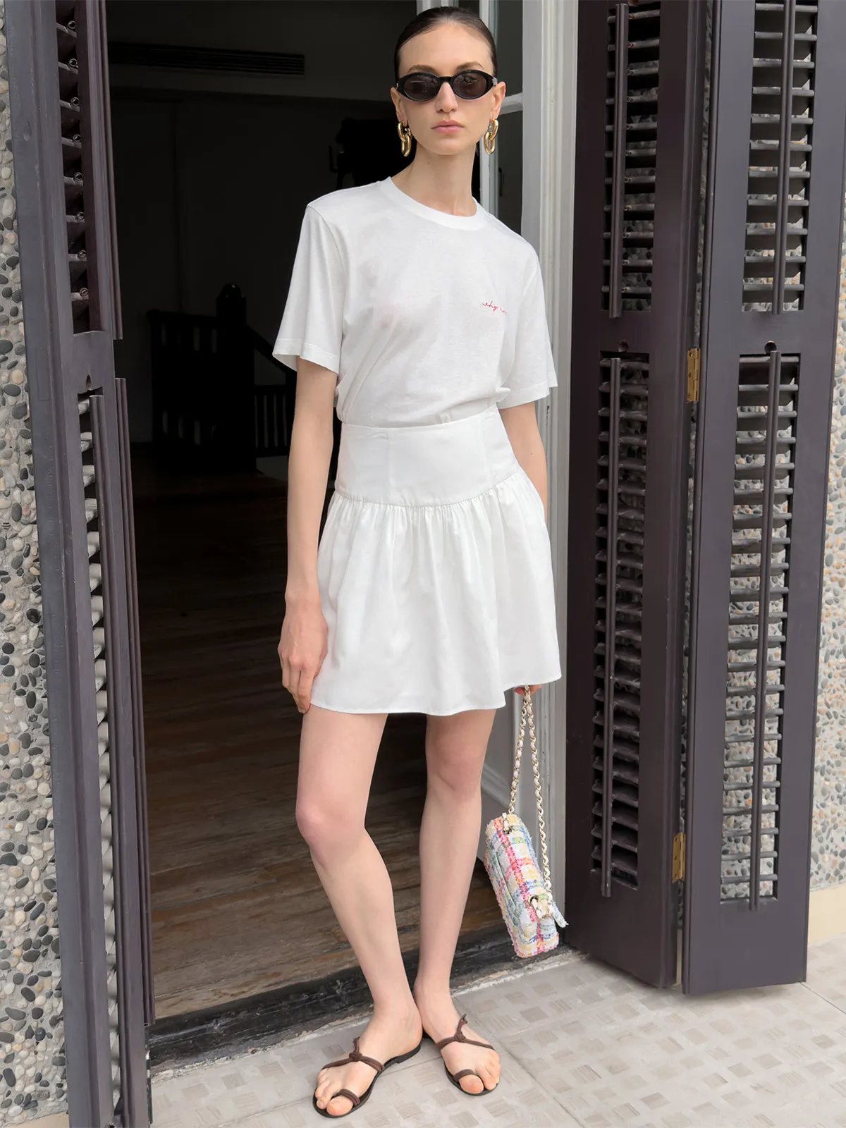 Model standing in doorway wearing white mini skirt with white tee, sunglasses, and colorful chain bag