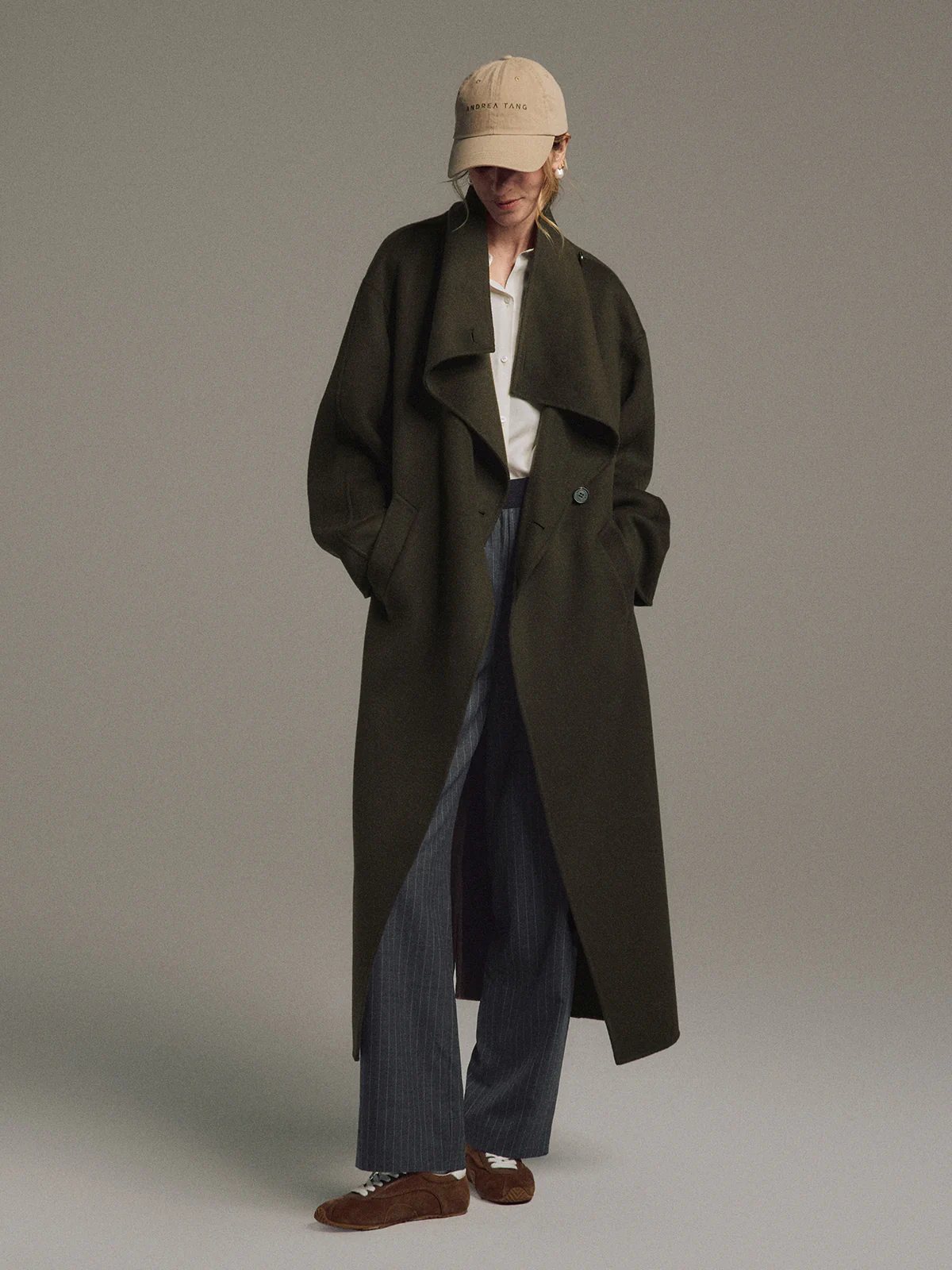 Model wearing the Olive TANSY long coat with hands in pockets, paired with striped trousers and a cap, side-front pose.