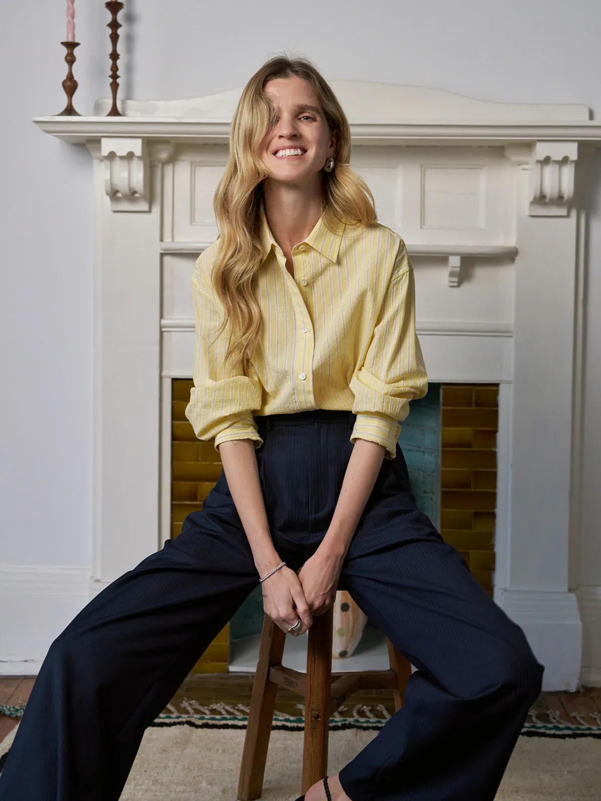Smiling model in buttercup shirt and navy wide-leg trousers seated on stool before white fireplace