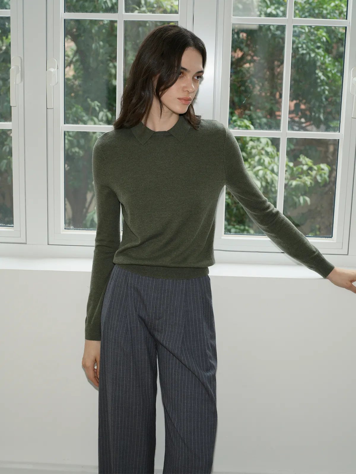 Model in olive green LOUANE jumper by large window with green foliage outside
