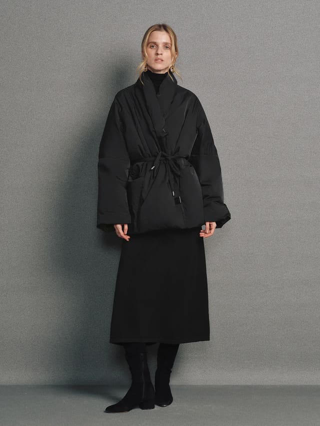 Model wears the Black OPHELIA relaxed wrap-style down coat with wide sleeves, tied waist, and hands in pockets.