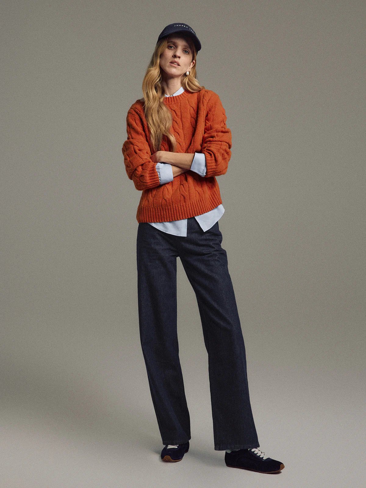 Model in an orange cable knit jumper layered over a shirt, wearing dark jeans and a cap with arms crossed.