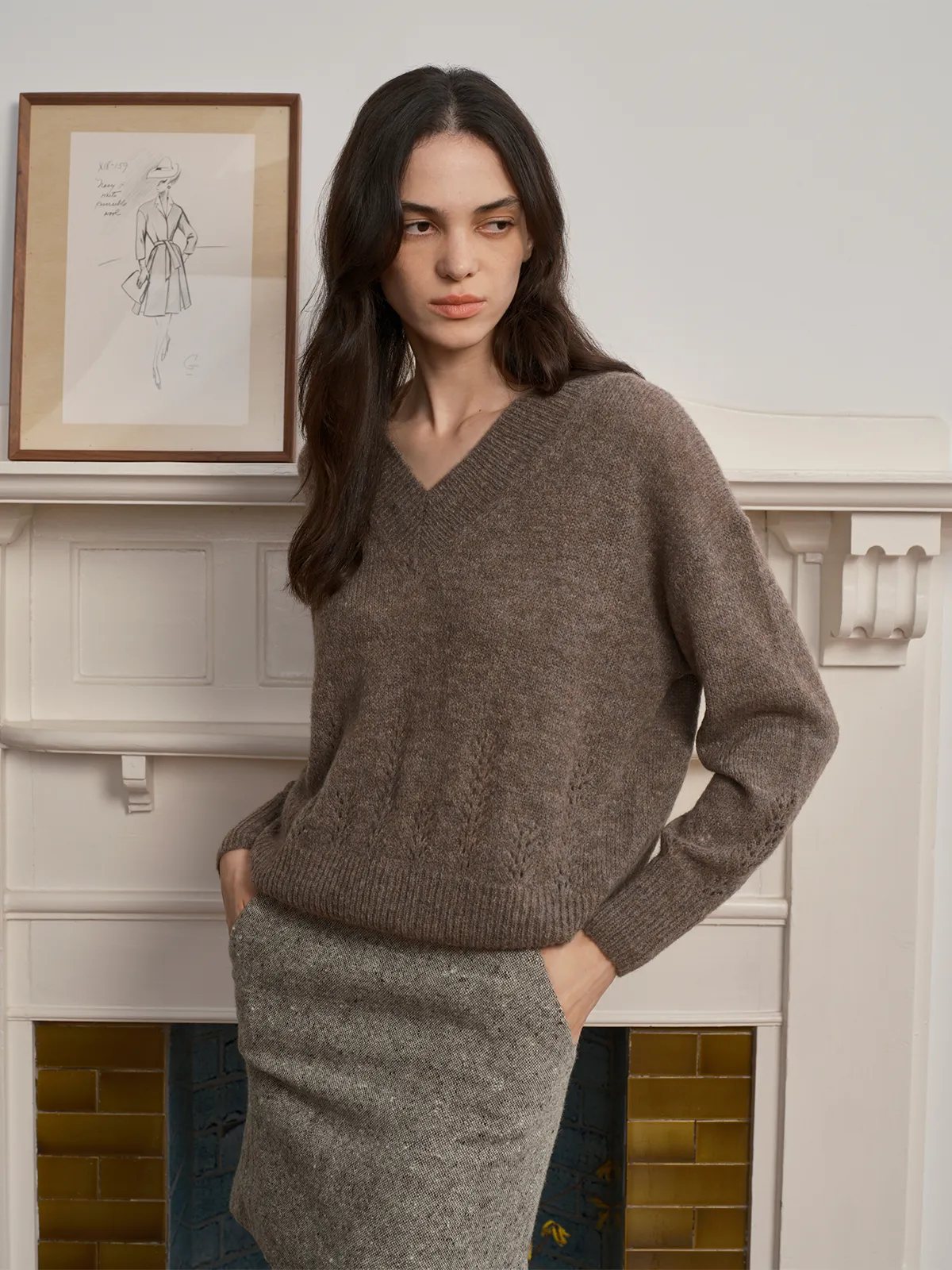 Model in taupe AIRY Jumper with grey tweed pencil skirt, hands in pockets, beside fireplace with fashion sketch