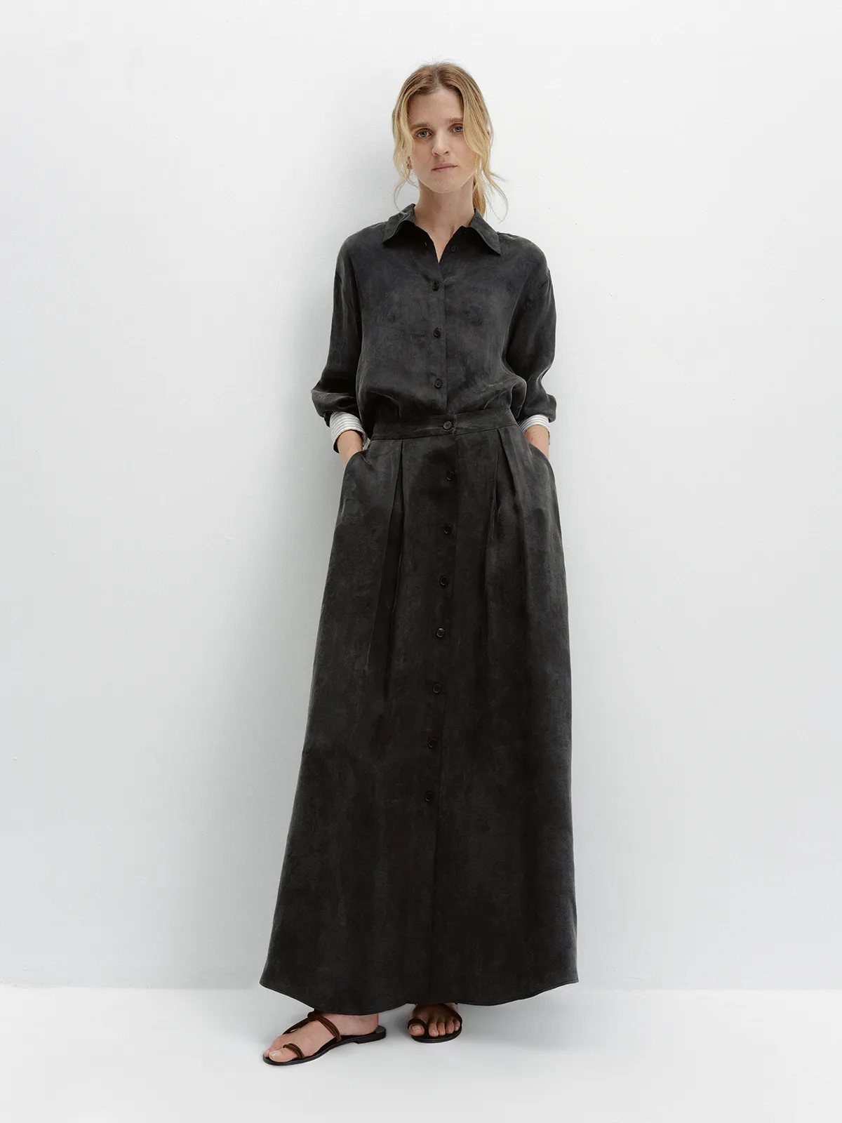 Model wearing black NOBU Maxi Skirt with matching shirt, hands in pockets, minimalist white background