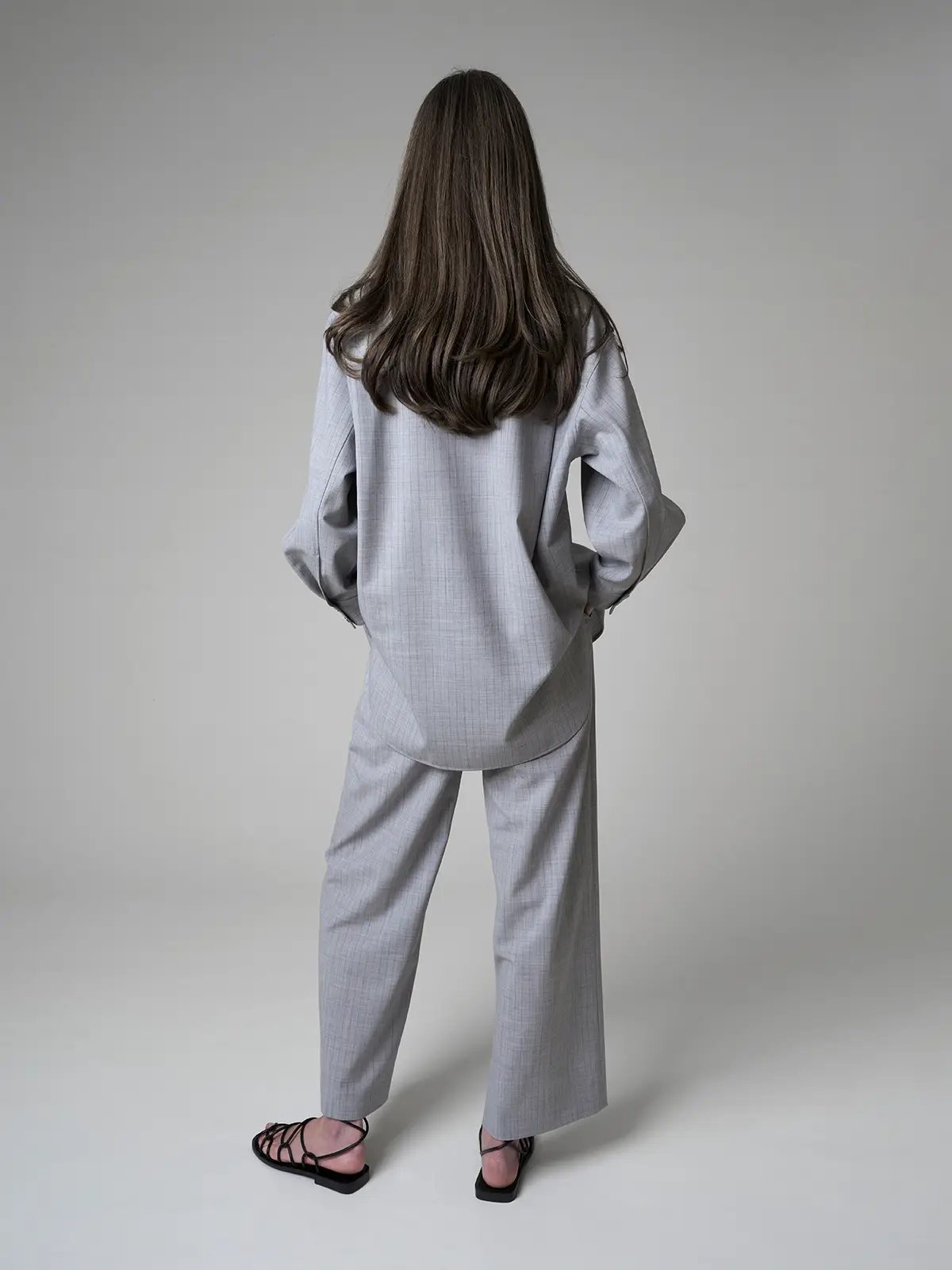 Rear view of model wearing the grey striped loose wool shirt and matching trousers, showing relaxed drape and back hem detail.