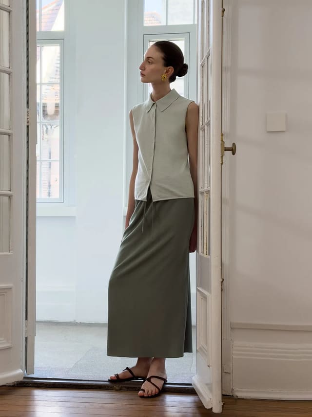 Model standing in doorway wearing MARGOT knit shirt with olive maxi skirt and black strappy sandals