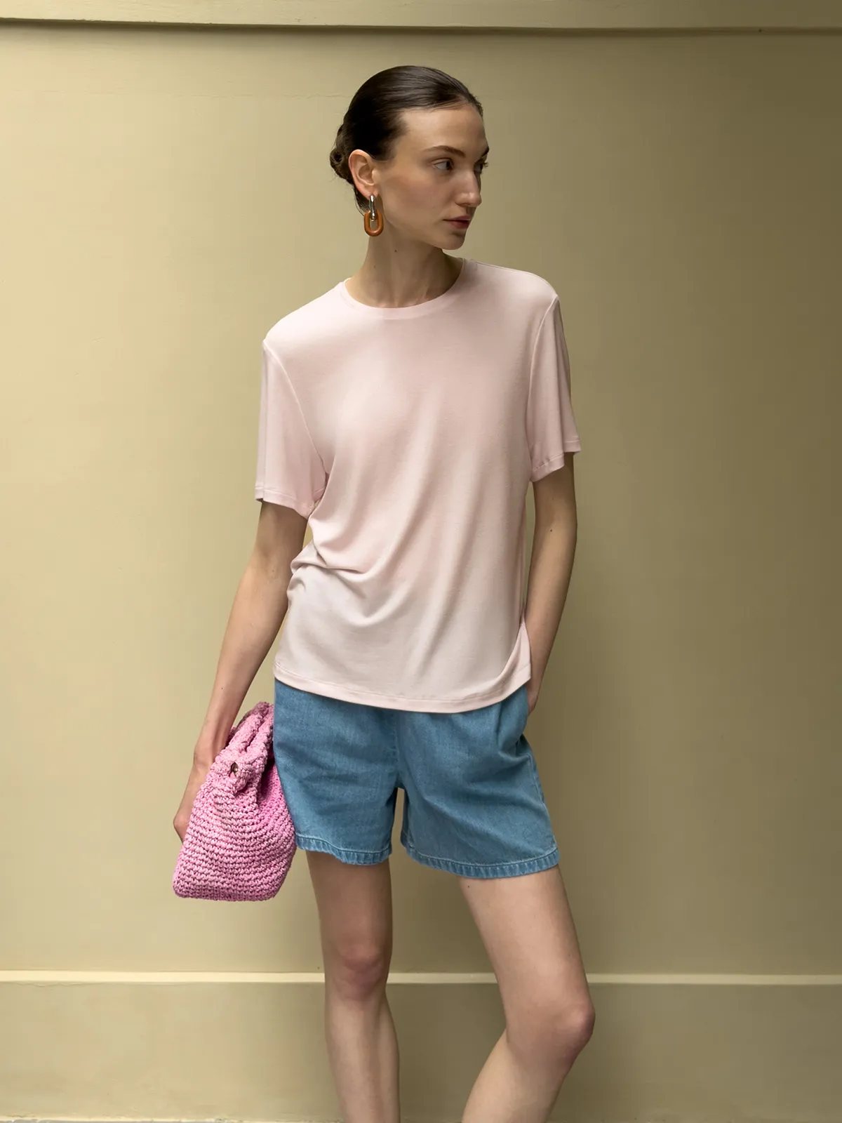 Model in blush pink tee and WAYLON denim shorts holding pink crocheted clutch against neutral wall
