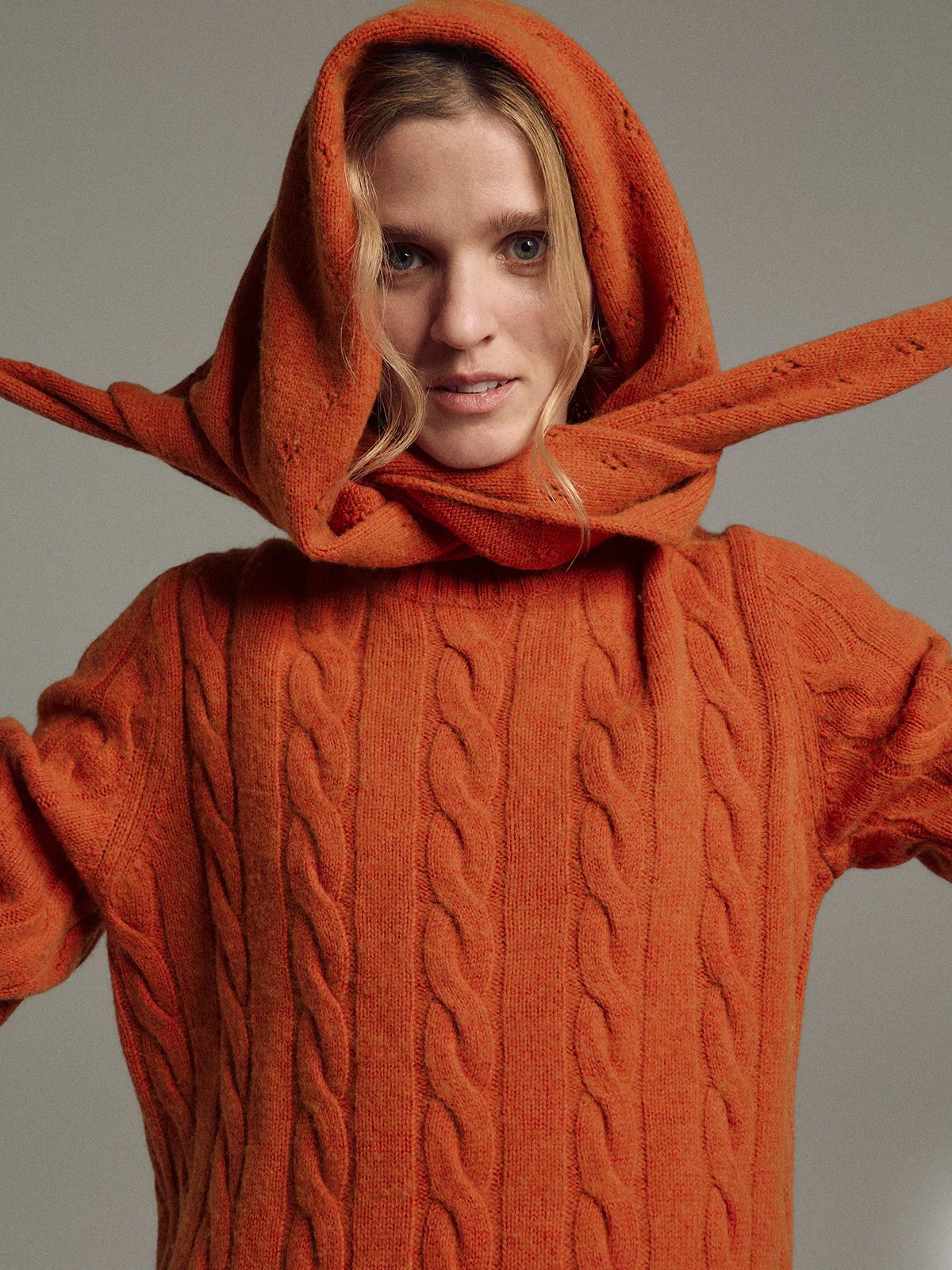 Model in orange yak wool triangle scarf styled as a hood, framing the face over a cable-knit sweater.