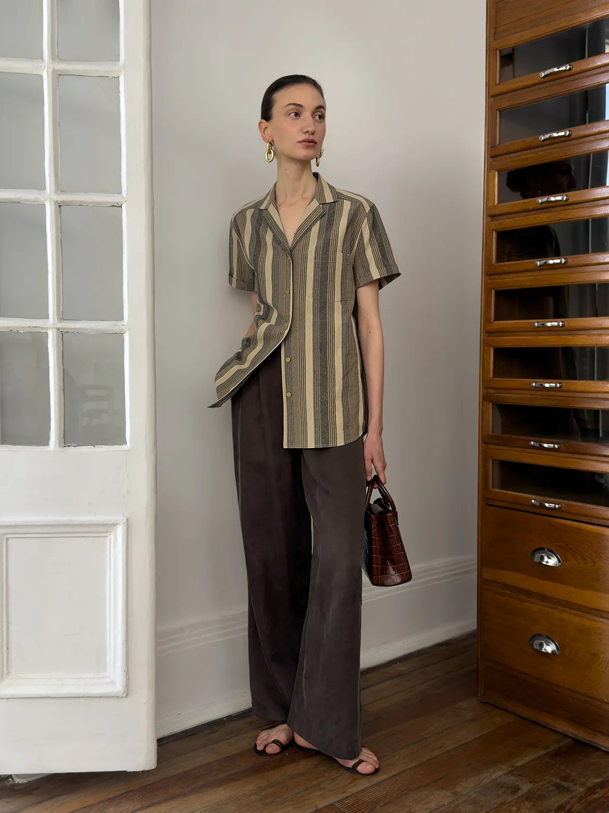 Model wearing REAGAN striped shirt with brown wide-leg trousers and croc-embossed bag in vintage room setting