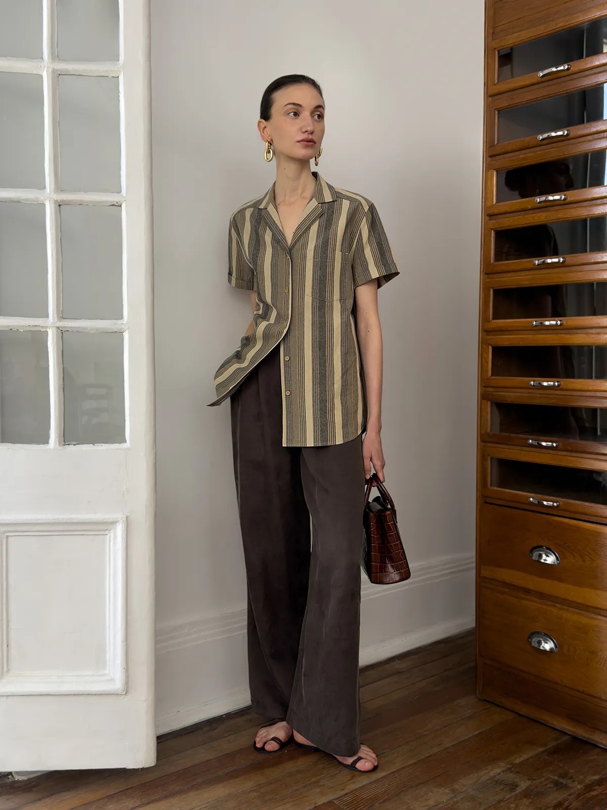 Model wearing REAGAN striped shirt with brown wide-leg trousers and croc-embossed bag in vintage room setting