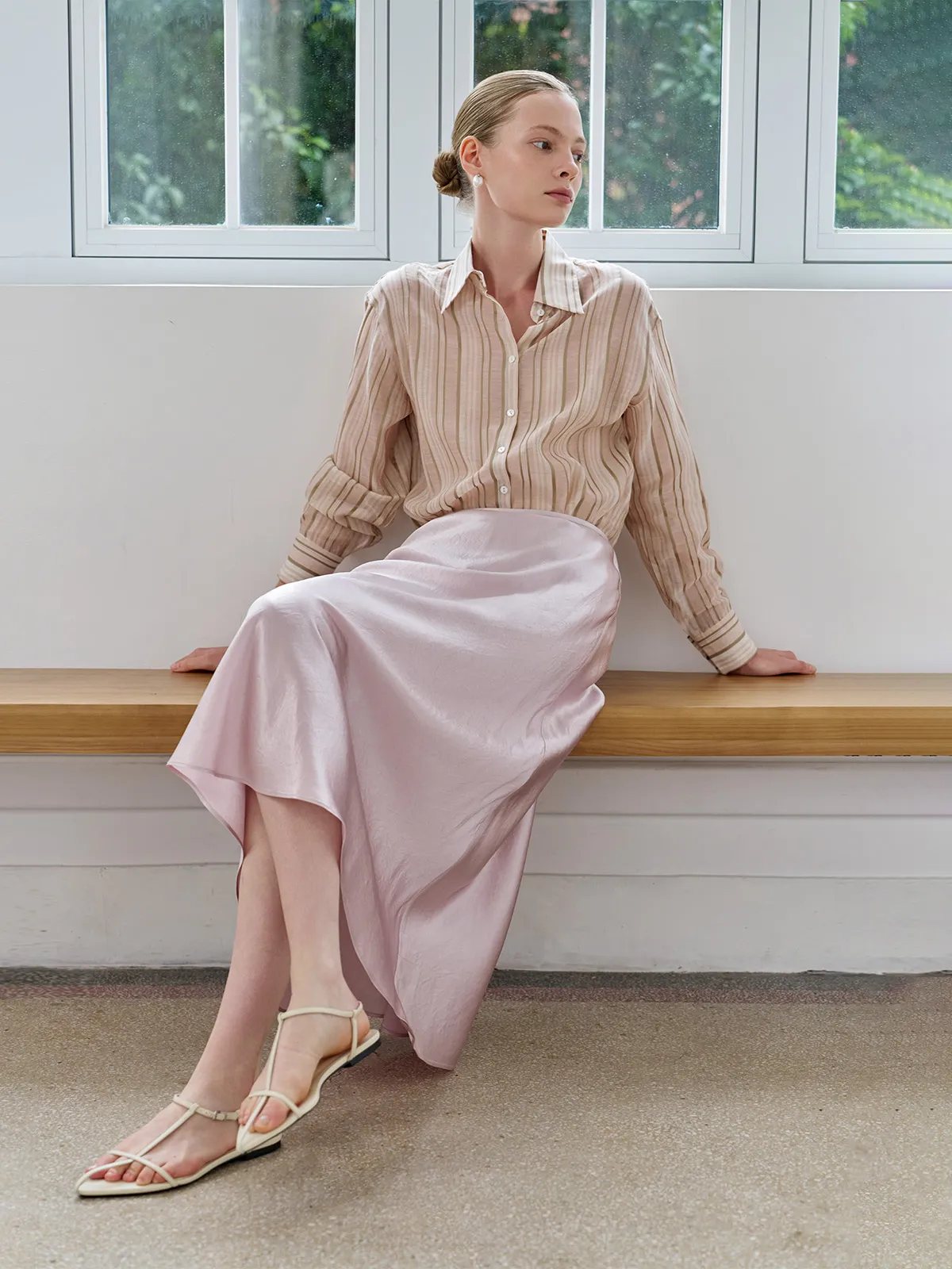 Model seated on wooden bench wearing SHELL skirt with beige striped shirt and strappy sandals, side profile