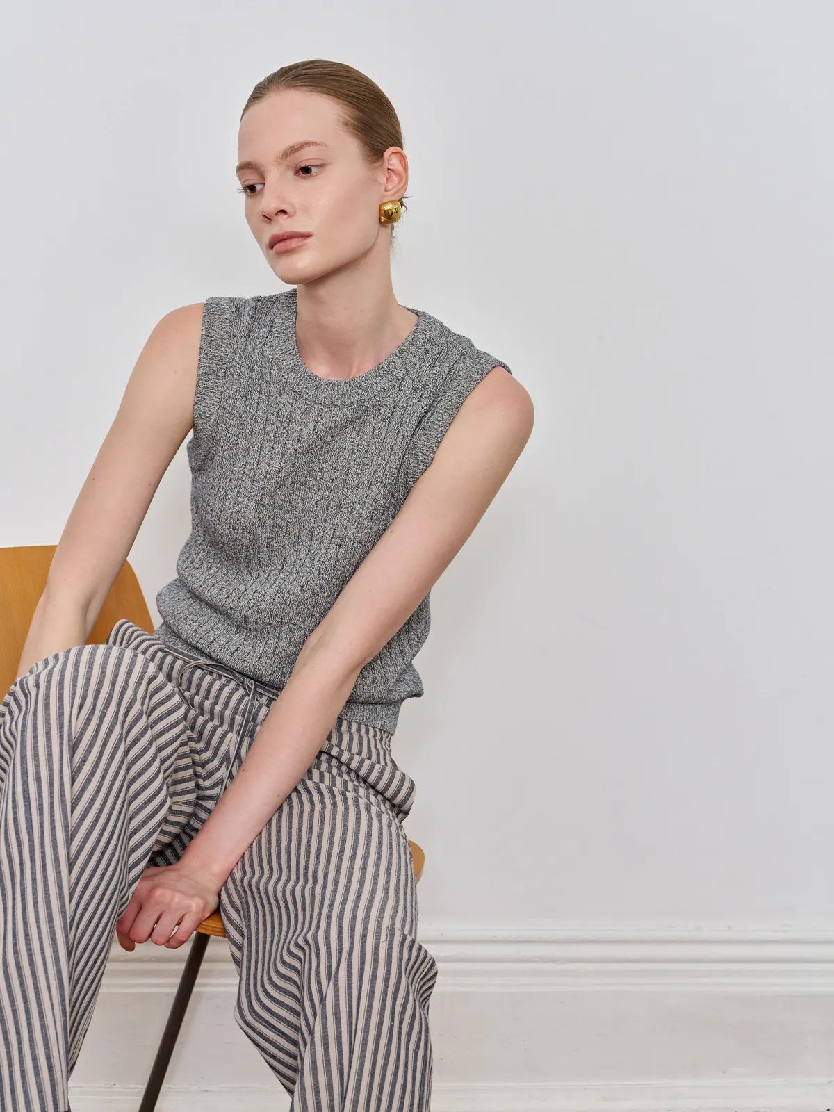 Model in grey MILLIE tank top seated sideways on chair, highlighting shoulder drape and ribbed hemline — minimalist fashion photography