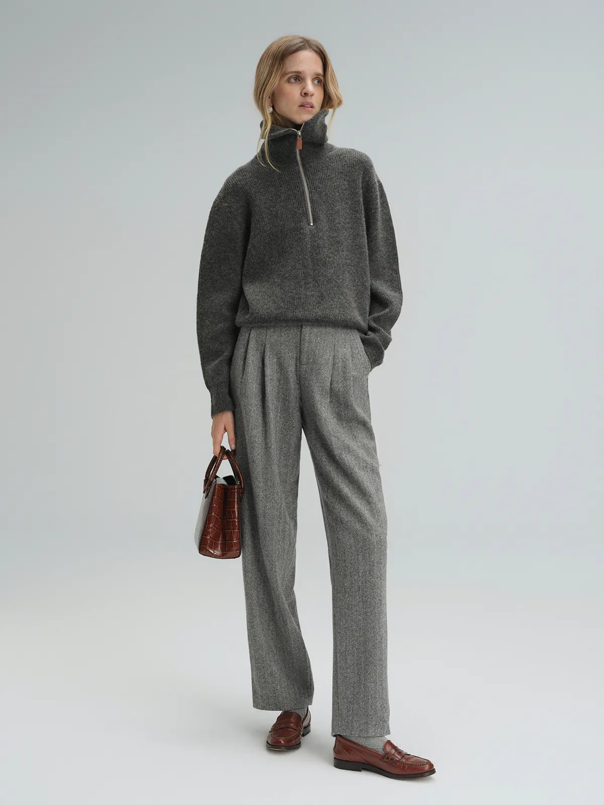Side pose of model wearing Gray wool herringbone tapered trousers with a gray zip-neck sweater and loafers.