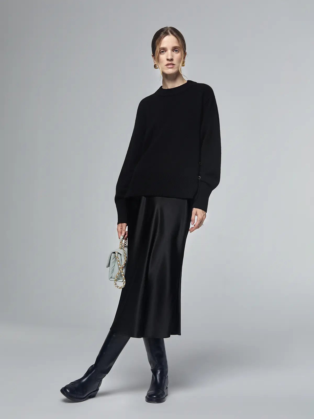 Model wearing a black merino wool sweater with metal buttons and ribbed hem, styled with a black skirt and boots.