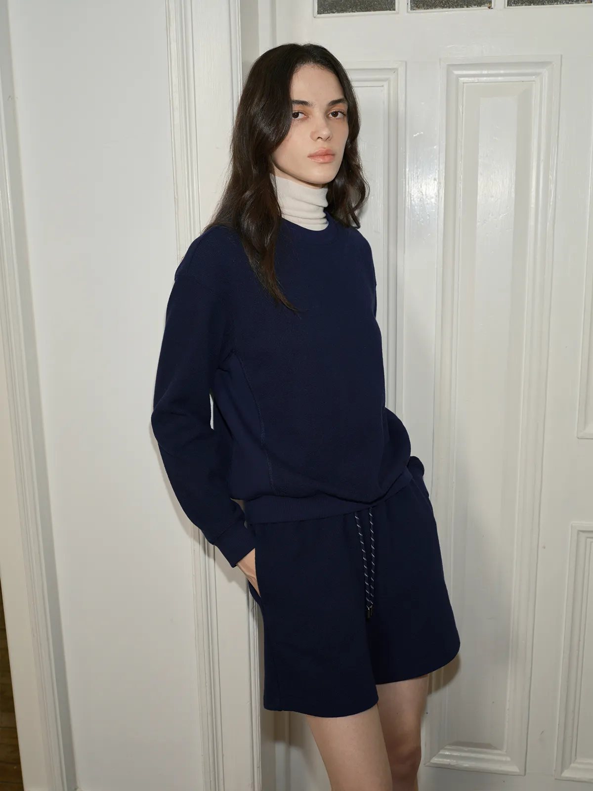 Model in navy ARLO terry short sweatpants paired with navy crewneck and cream turtleneck, hands in pockets against white door