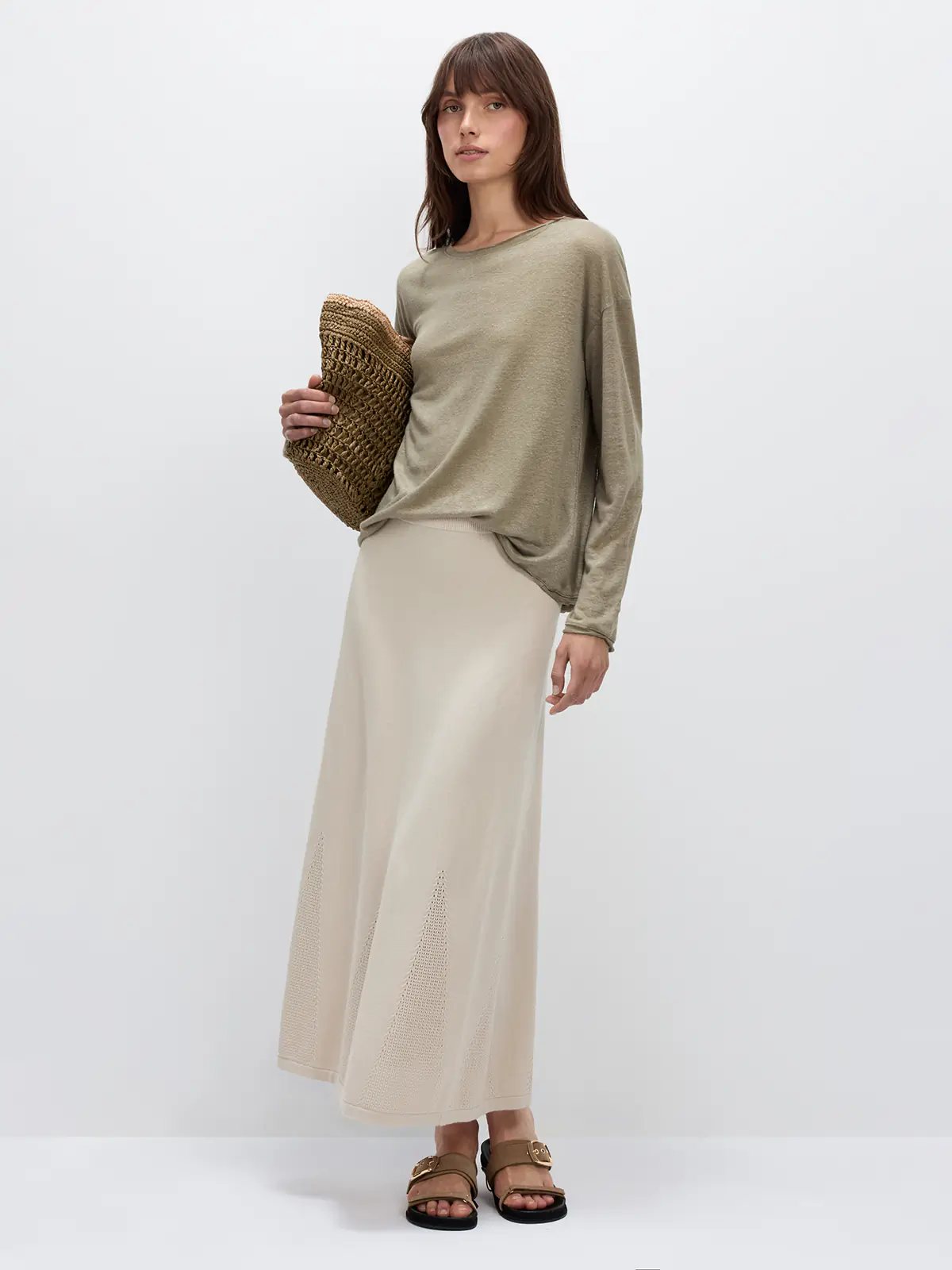Model in a grayish green lightweight long-sleeve linen t-shirt with dropped shoulders, styled with a midi skirt and woven bag.