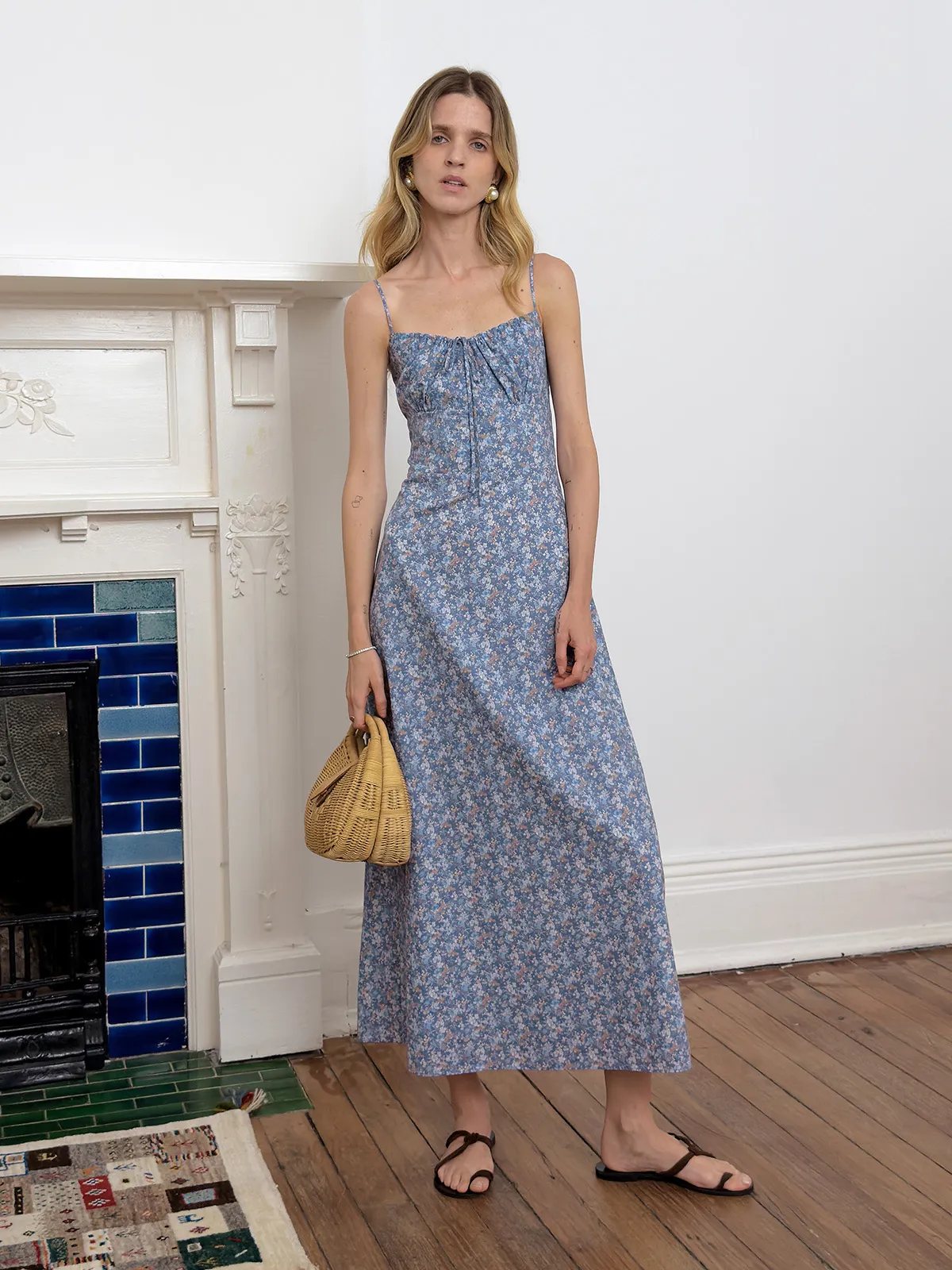 Model in blue floral PIPPA dress holding woven straw bag, standing beside ornate fireplace with blue tiles