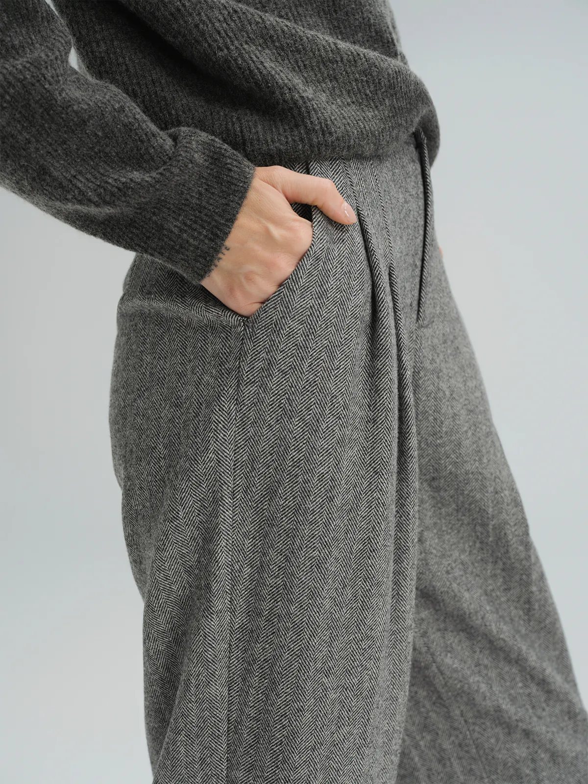 Close-up of hand in pocket of Gray wool herringbone tapered trousers, showing herringbone texture and pleat detail.