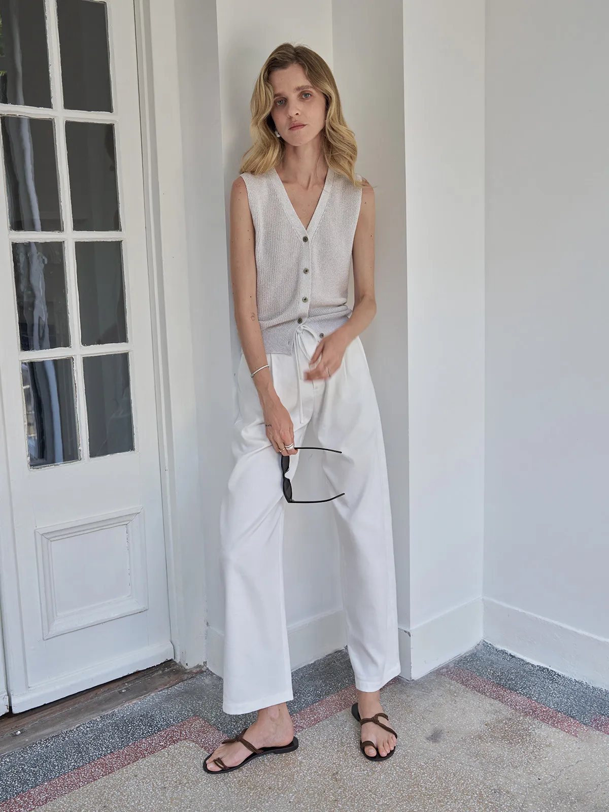 Model leaning against wall in white LEIGHTON trousers, holding sunglasses, showcasing side profile and waist tie