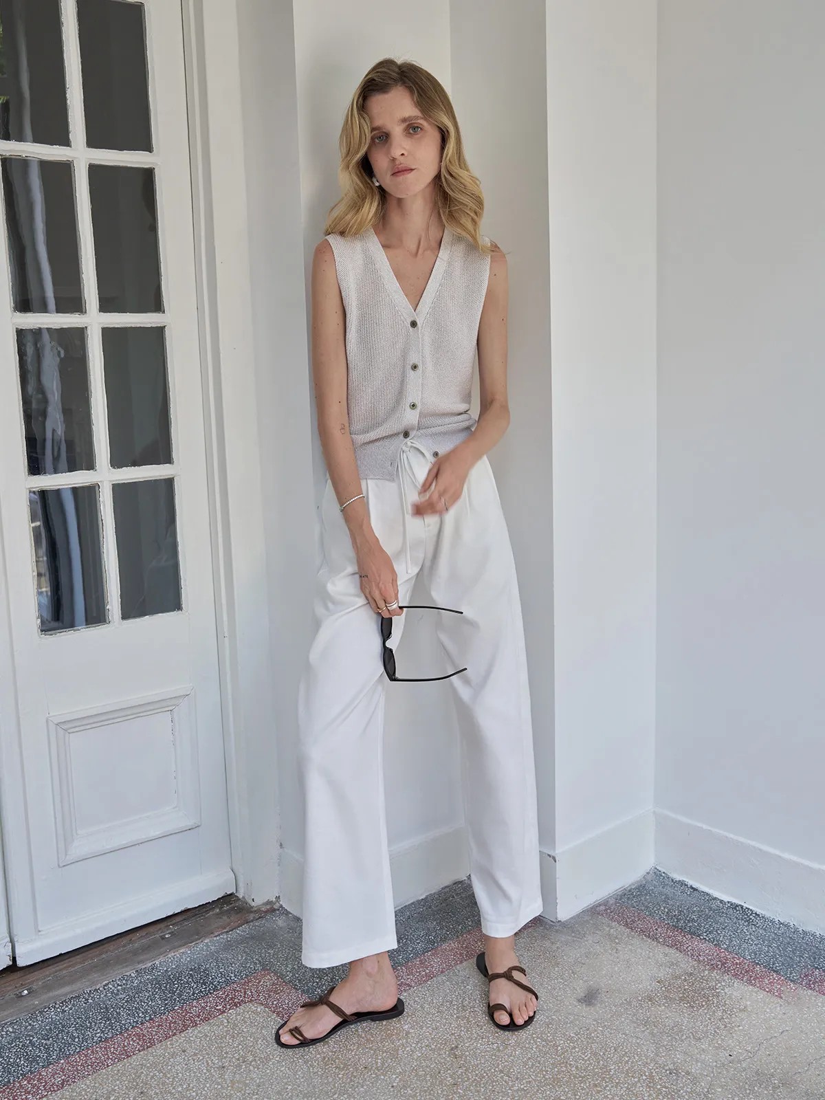 Model leaning against wall in white LEIGHTON trousers, holding sunglasses, showcasing side profile and waist tie