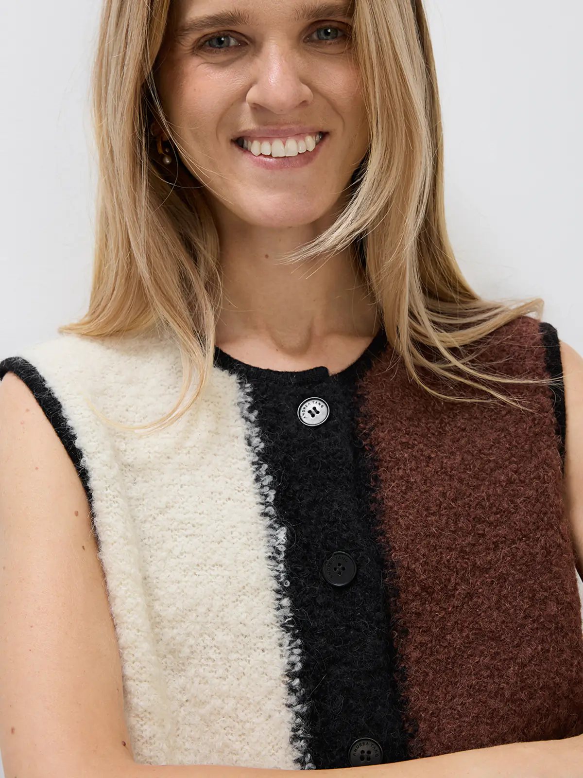 Model stands front-facing in a sleeveless Brown/White/Black colorblock knitted vest with buttons and a pocket, paired with brown trousers and holding a brown bag.