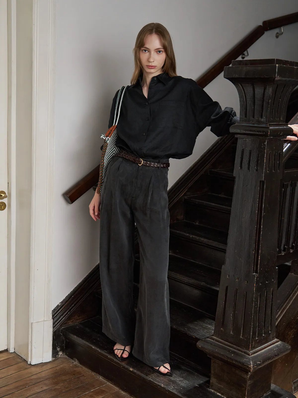 Model on vintage wooden staircase wearing black linen BREATH shirt tucked into charcoal wide-leg trousers with braided leather belt and woven bag