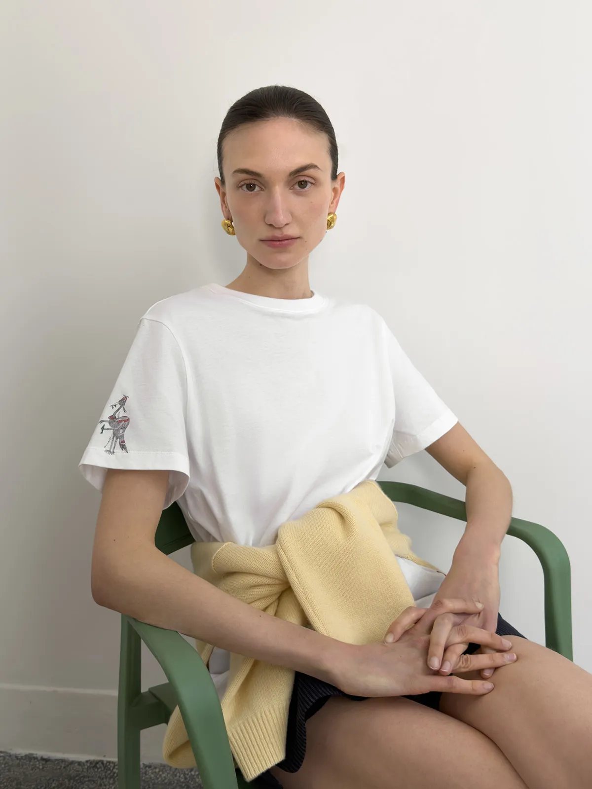 Front portrait of model seated in green chair wearing white EDWARD t-shirt with sleeve crane embroidery, yellow knit draped at waist — quiet luxury aesthetic