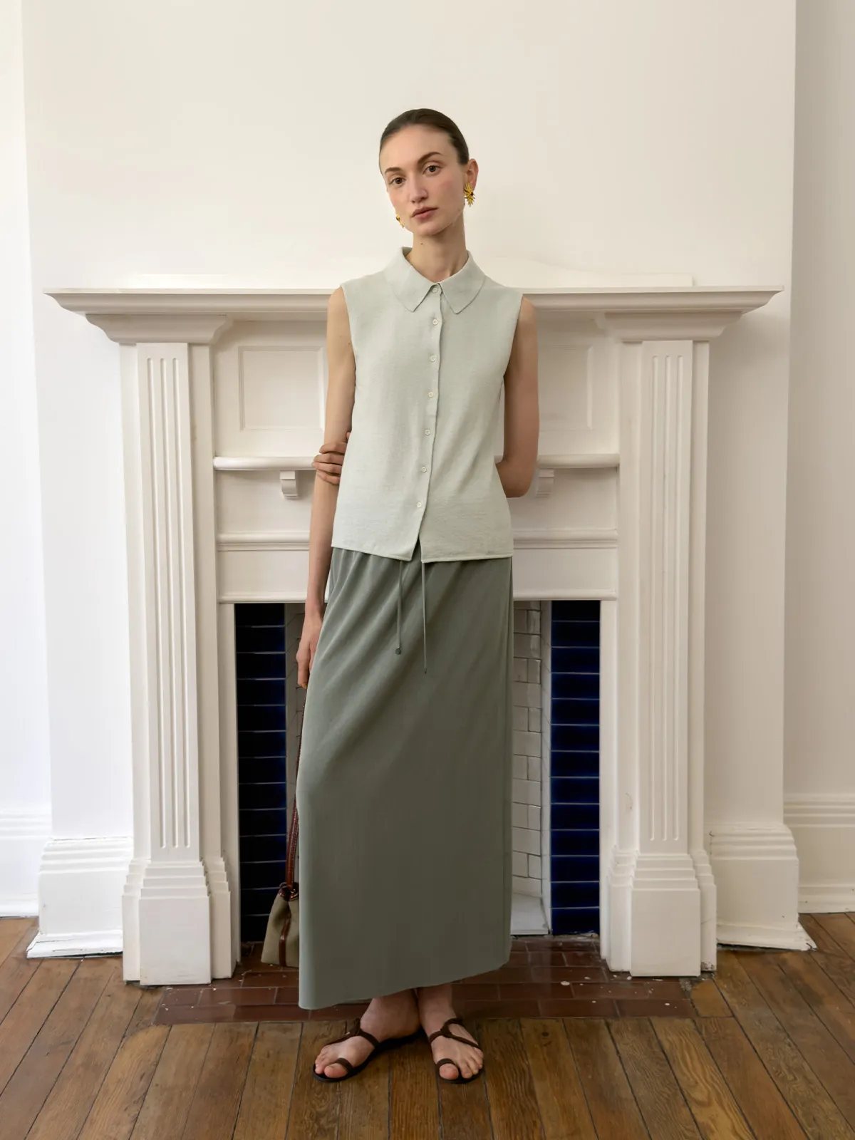 Model standing in front of white fireplace wearing MARGOT sleeveless shirt with olive green maxi skirt and sandals