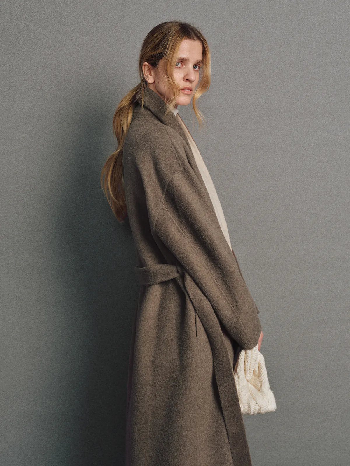 Model wearing brown two-tone reversible notched-collar coat open over turtleneck, holding clutch.