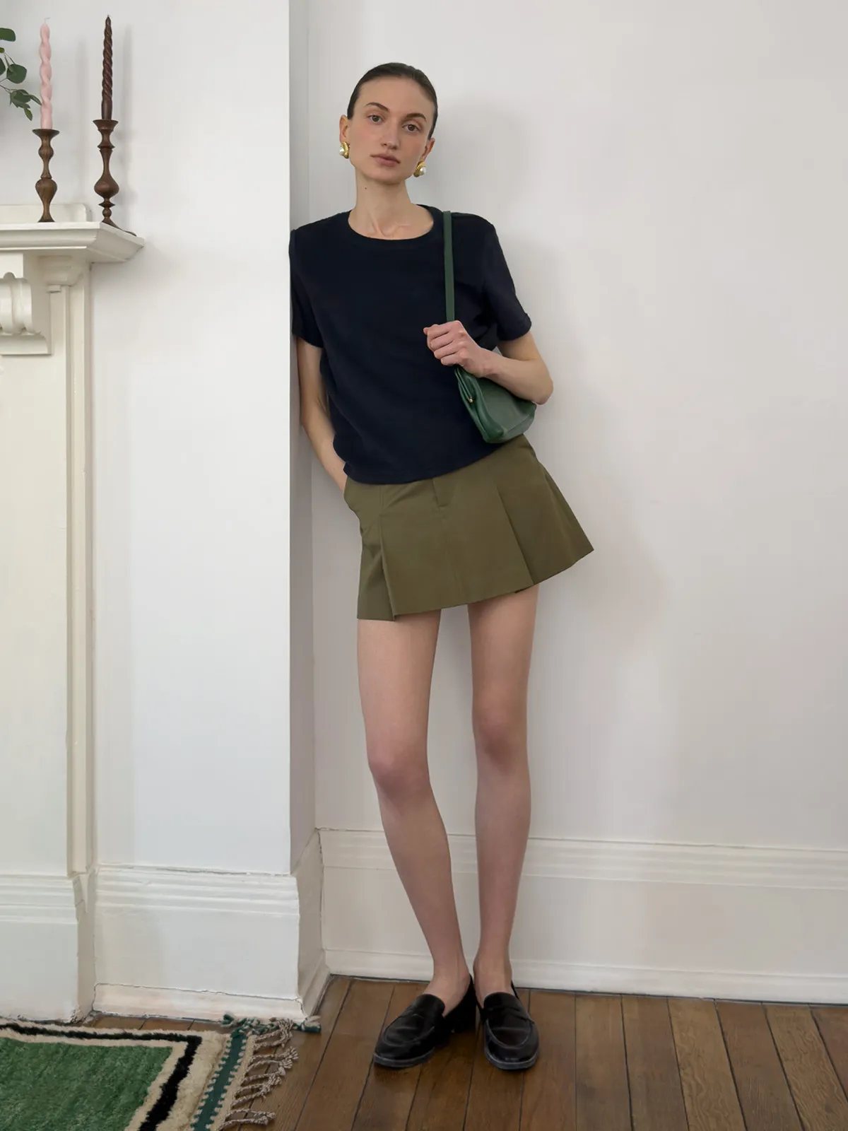 Andrea Tang LANE Terry short sleeve t-shirt in navy paired with olive pleated mini skirt and green leather bag, minimalist styling against white interior wall