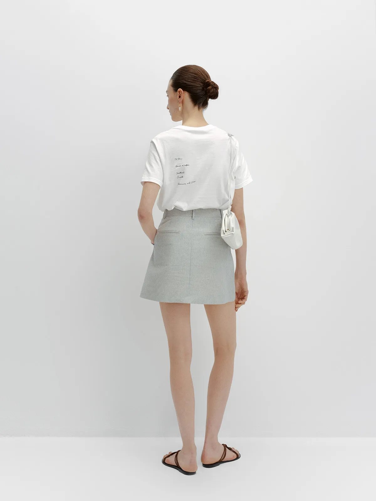 Back view of model wearing white embroidered t-shirt with grey striped mini skirt and white shoulder bag