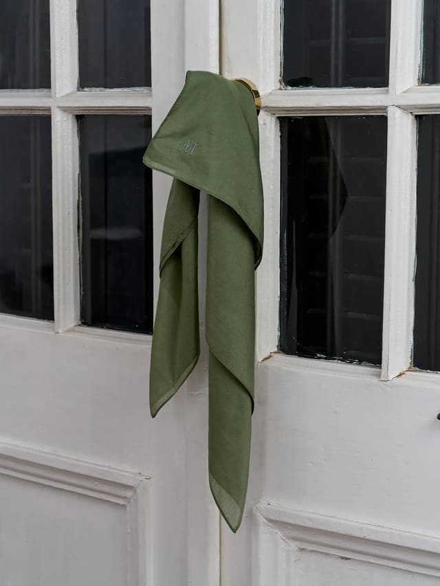 Olive green TIANA silk cotton square scarf with embroidered AT monogram hanging on white door handle, minimalist aesthetic