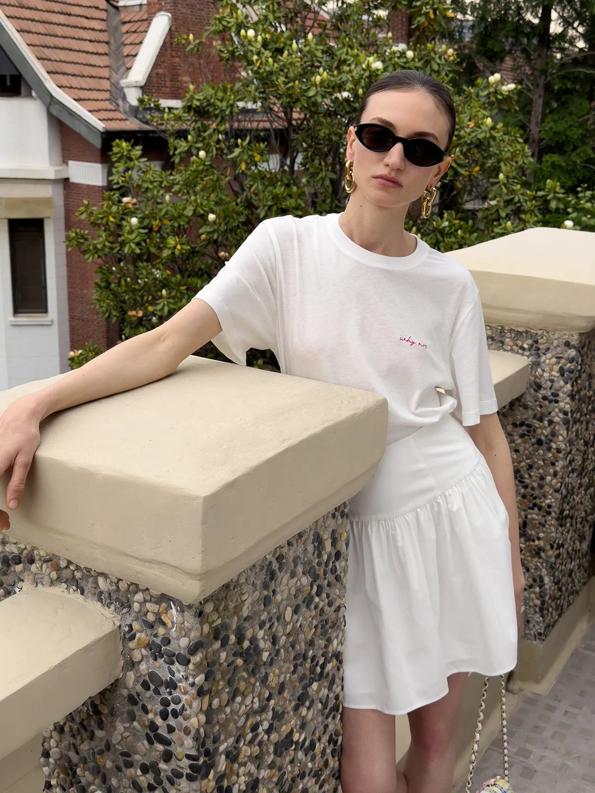 Model wearing white relaxed t-shirt with red 'why not' embroidery, paired with white tiered skirt, outdoors on balcony